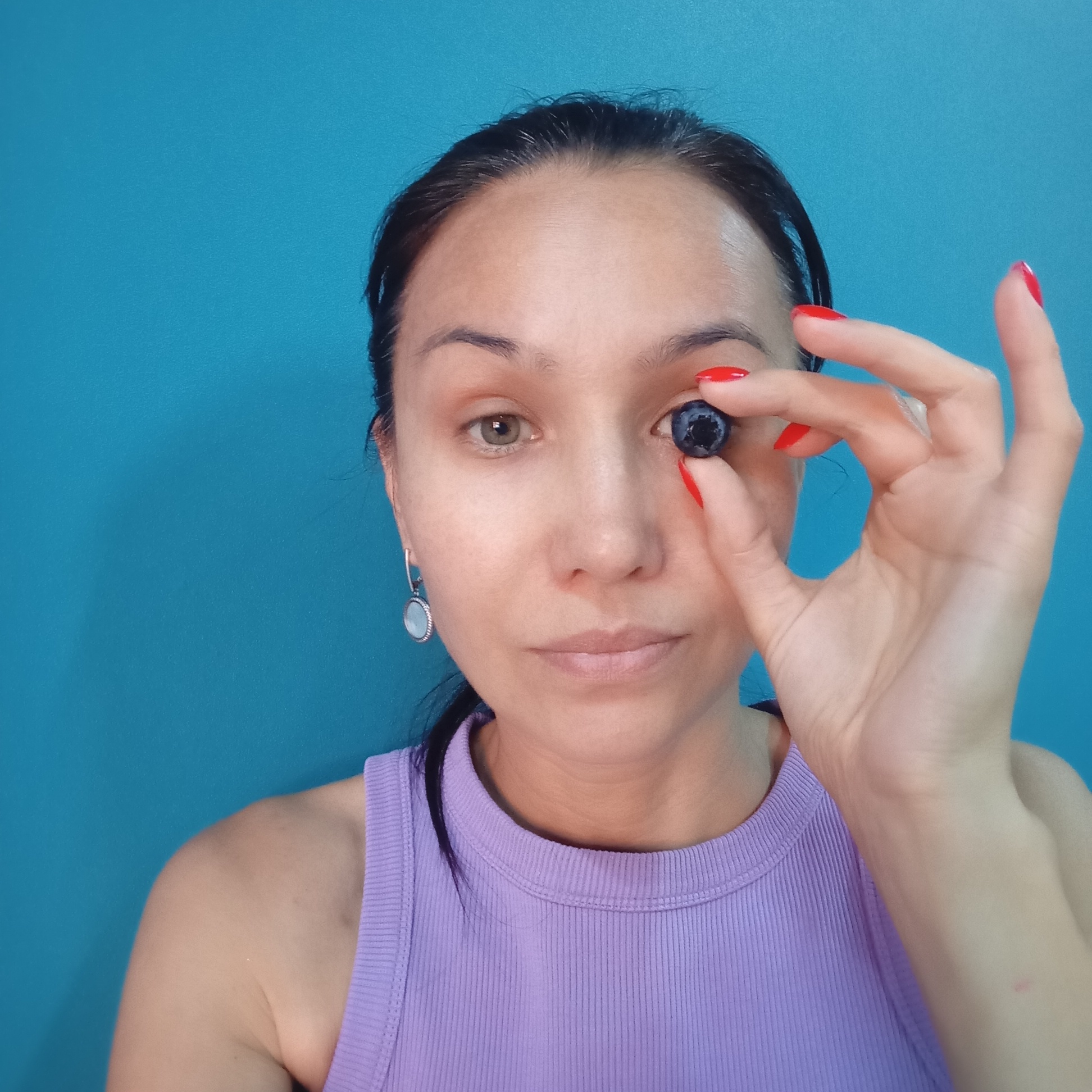 A medium close-up shot captures a woman against a solid blue background. She is holding a single blueberry in front of her right eye with her manicured red-nailed fingers, partially obscuring her pupil. Her left eye is visible and appears to be a different color from her right, which is covered by the blueberry. She wears a purple ribbed tank top and a silver earring with a light-colored circular pendant in her left ear. Her hair is dark and pulled back. The lighting is even, and the image appears to be a selfie. The location in the city of Pervomaysky, Russia, is not determinable from the visual information provided.