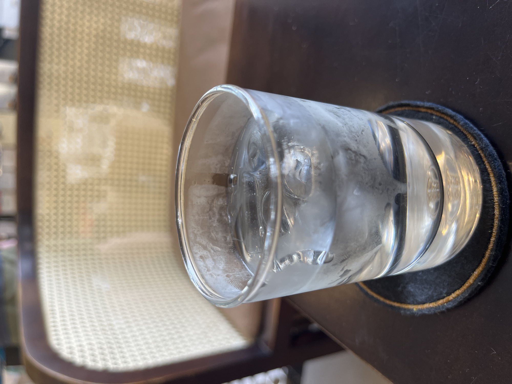 A close-up, angled shot features a glass of water with ice sitting on a black coaster with a gold trim, placed on a dark wooden table. Condensation covers the exterior of the glass. To the left of the glass is a chair with a woven seat and dark wooden frame. The background is blurred, but suggests an indoor setting. The location is Nha Trang Ward, Vietnam.