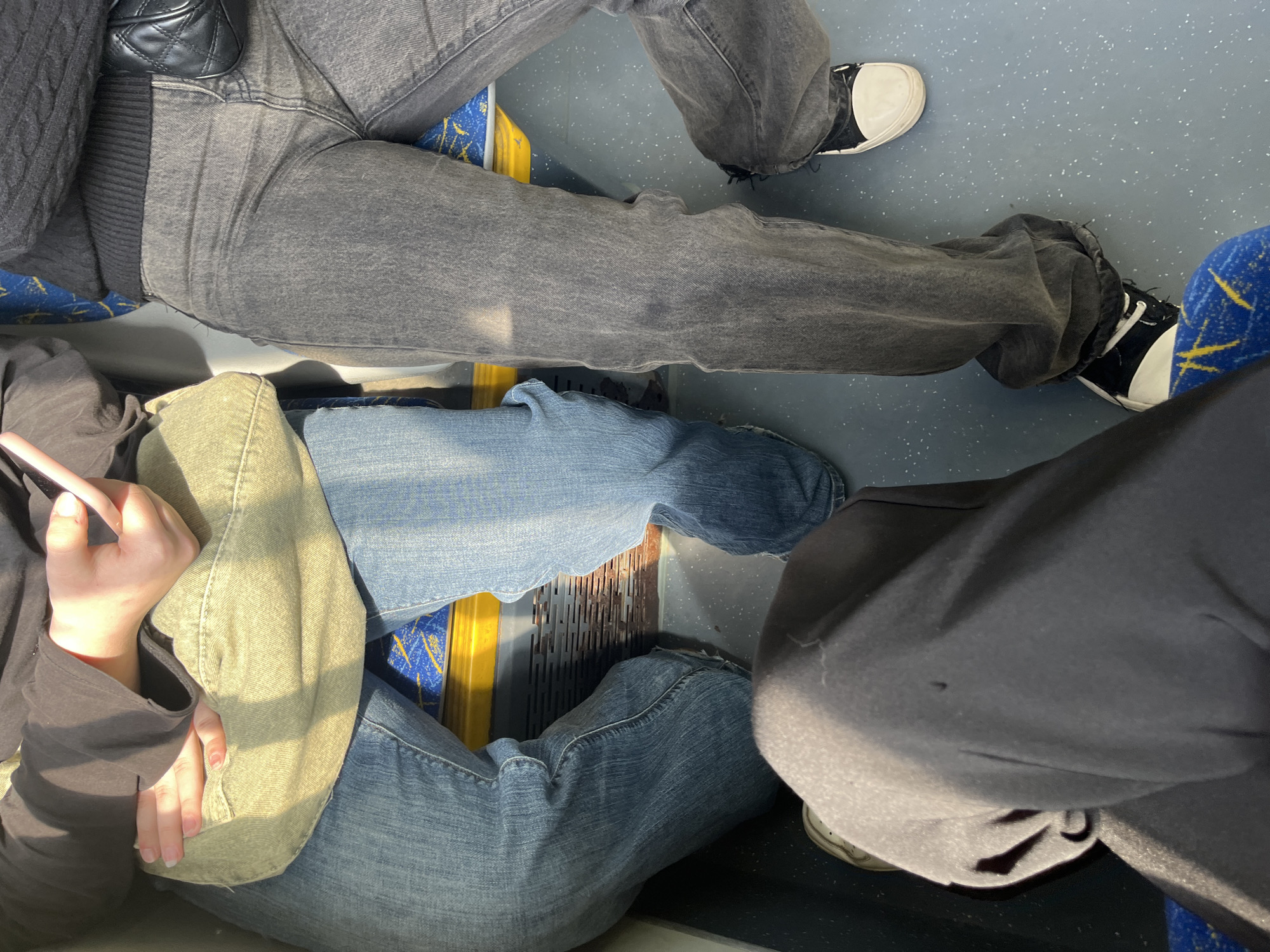 An elevated, downward-angled shot captures the lower bodies of four individuals seated inside what appears to be a public transport vehicle in Saint Petersburg, Russia. Bright sunlight illuminates the scene, suggesting it is daytime.
In the upper portion of the frame, two people are visible. One wears grey jeans and a dark, possibly knitted, top, with a black quilted shoulder bag resting on their lap. Adjacent to them are the legs of another person, also in grey jeans and black-and-white high-top sneakers, with their right foot near a blue seat featuring a yellow, abstract pattern.
Towards the bottom left, a person is seen wearing blue jeans and a light green or khaki jacket over a dark long-sleeved top. Their hands are clasped, holding a light-colored object, possibly a phone. Their legs are positioned near a yellow bar and a metallic grate on the floor, which is grey with white speckles.
To the bottom right, the legs of a fourth person are visible, dressed in dark (likely black) trousers or sweatpants. A white-soled shoe is partially seen. The blue and yellow patterned seats are a prominent feature of the interior.
The overall scene depicts passengers traveling within a vehicle, with no specific activity beyond sitting. No visible text is present in the image.
