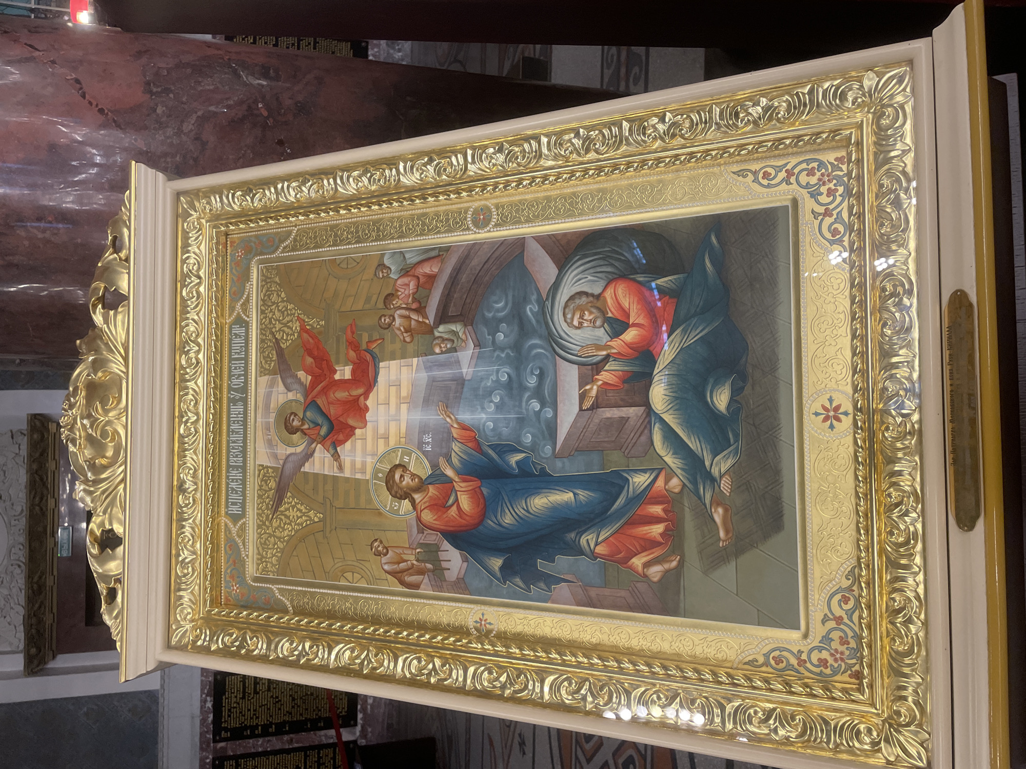The image depicts an icon, likely displayed in the city of Kronstadt, Russia, given the inscription at the bottom. The icon is housed in an ornate, gilded frame with decorative motifs. The central scene of the icon illustrates the biblical event of Jesus Christ raising Lazarus from the dead.
Jesus, depicted with a halo and wearing a blue robe over a red garment, stands to the left of the frame. He gestures with his right hand towards Lazarus, who is shown emerging from a tomb or sarcophagus. Lazarus, with a beard and wearing an orange robe, is depicted with his arms raised and his body partially uncovered, suggesting his resurrection from death.
Above Jesus, an angel with wings and a halo flies down, with rays of light emanating from the heavenly realm. In the background, several figures are observing the scene from a stone structure that appears to be a bridge or walkway. One figure, a man with a beard and a grey garment, is seated near Lazarus's tomb.
The environment depicted in the icon is stylized and symbolic, with a blue, wavy representation of water in the foreground and stone architectural elements. The background features a golden sky or aura. The overall style is consistent with traditional Eastern Orthodox iconography. The inscription at the bottom of the icon likely provides further details about its provenance or subject matter.