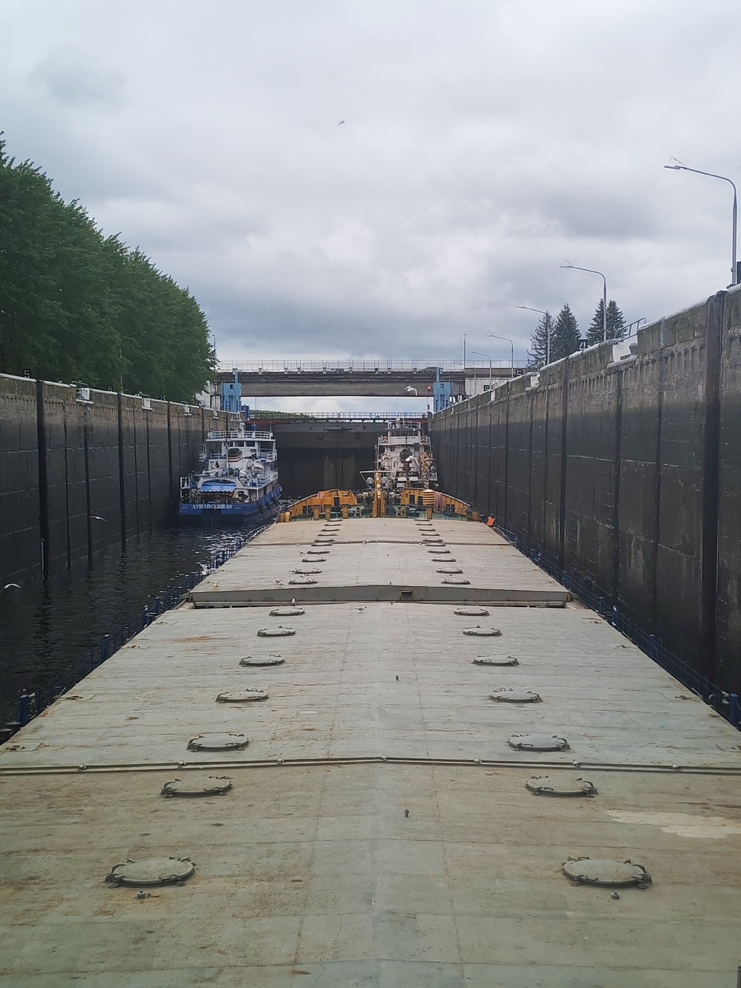 The image shows a barge in a lock on a canal in the city of Unknown. The barge is surrounded by high concrete walls, with trees and streetlights visible on the upper edges of the walls. There is a bridge spanning the canal. A smaller boat is visible behind the barge. The barge's deck is covered with numerous circular hatches. The sky is overcast.