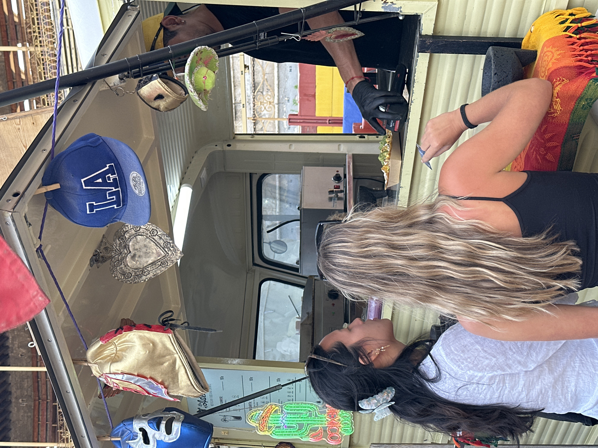 The image depicts an outdoor food truck or stand, presumably in the city of Unknown, Unknown. The stand is decorated with a Mexican theme. **People:** * **Vendor:** A male individual is partially visible on the right, wearing a black t-shirt and a yellow cap. His right hand, covered by a black glove, is seen interacting with the counter, possibly preparing or serving food. A red string bracelet is on his left wrist. * **Customer 1:** A female with long dark hair, styled with a light blue butterfly-shaped claw clip, stands on the left. She wears a white, textured, long-sleeved shirt and earrings. She is looking towards the food counter. * **Customer 2:** A female with long, wavy blonde or light brown hair is positioned to the right of Customer 1, with her back mostly facing the camera. She wears a black tank top. Her right arm rests on a colorful textile, and she holds a dark blue object, possibly a mobile phone or utensil, in her hand. **Objects and Environment:** * **Food Stand:** A light yellow/beige colored mobile food stand is the central focus, with an open serving window. * **Decorations:** Hanging from a purple rope strung across the top interior of the stand are various items: a blue wrestling mask, a gold and red wrestling mask, a silver heart-shaped milagro ornament, and a blue "LA" baseball cap (Snapback brand). Further right, a small green sombrero with a pink and yellow band, adorned with a small yellow sphere, hangs. Another heart-shaped, flame-like milagro ornament hangs near the vendor's arm. A piece of red fabric is visible in the upper left. * **Signage:** Inside the stand, a light-colored menu board reads "Adelita's Mexican food and Drinks" with items and prices, including "Sunrise - 10c" and "Margarita - 10c." * **Neon Sign:** To the left of the menu board, a green neon sign shaped like a saguaro cactus wearing a sombrero illuminates, with "MEXICO" written in red neon below. * **Food:** A brown paper tray containing nachos or a similar dish is on the counter in front of the customers. * **Counter Details:** Inside the stand, kitchen equipment, including a silver appliance with knobs, is visible. Below the counter on the customer side, a black, rough-textured stone bowl (resembling a molcajete) rests on a surface. * **Textile:** A vibrant, multi-colored striped blanket or textile, featuring red, orange, yellow, and green stripes with