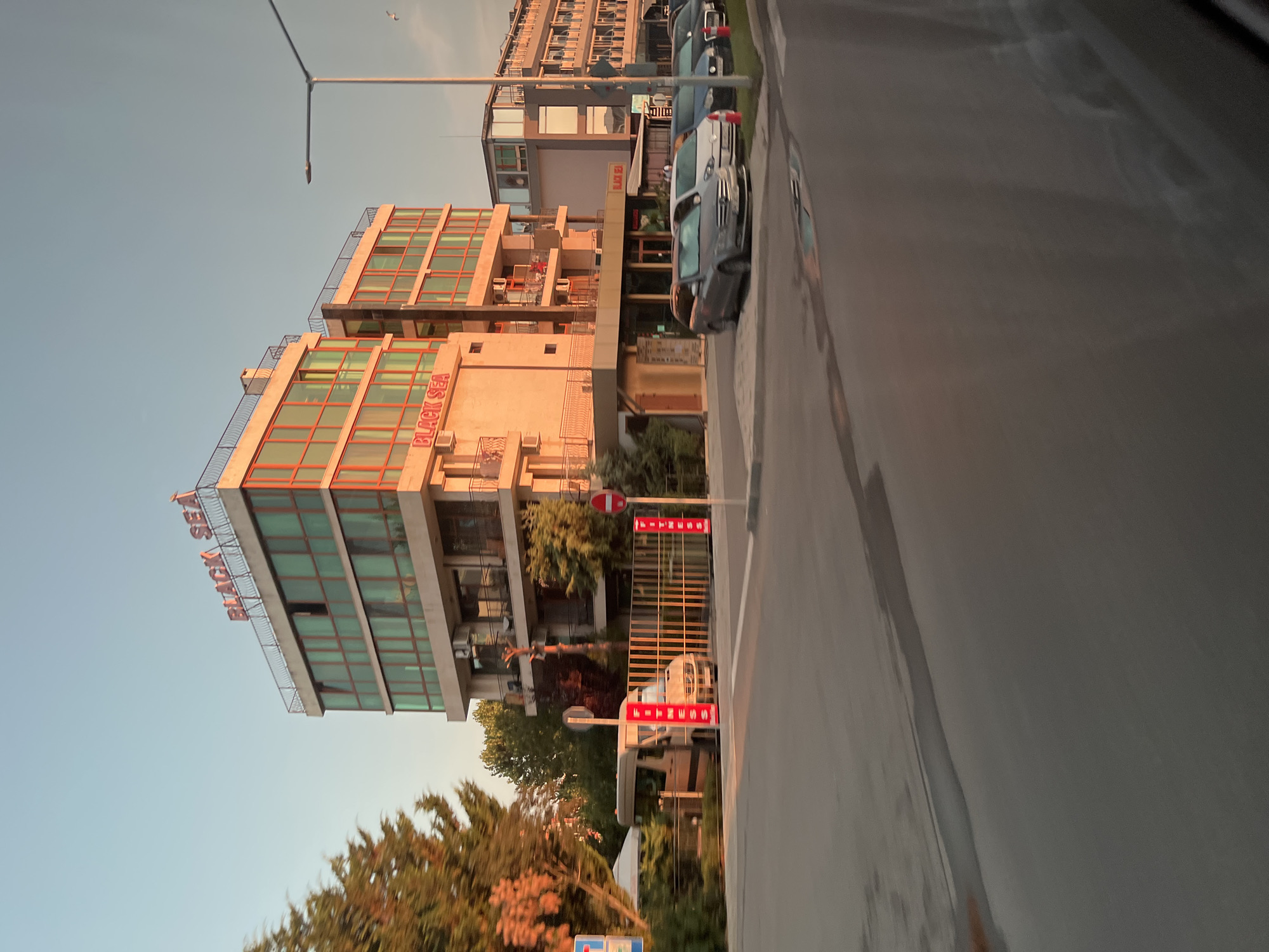 The image depicts a street scene in Nesebar, Bulgaria, featuring a multi-story building with a prominent "BLACK SEA" sign in red letters on its facade. The building has large, tinted windows framed in orange and a rooftop railing. Adjacent to it, other buildings are visible, suggesting a commercial or hotel area. Several cars are parked along the street, including a silver hatchback and a white hatchback. A light-colored van is parked near a fence with vertical yellow bars. Red and white striped poles with the word "FITNESS" are placed along the roadside. Trees with orange-tinged foliage are present on the left side of the image, and a street lamp stands on the right. The sky is clear and light blue, indicating daylight. The overall scene suggests a bright, possibly warm, environment.
