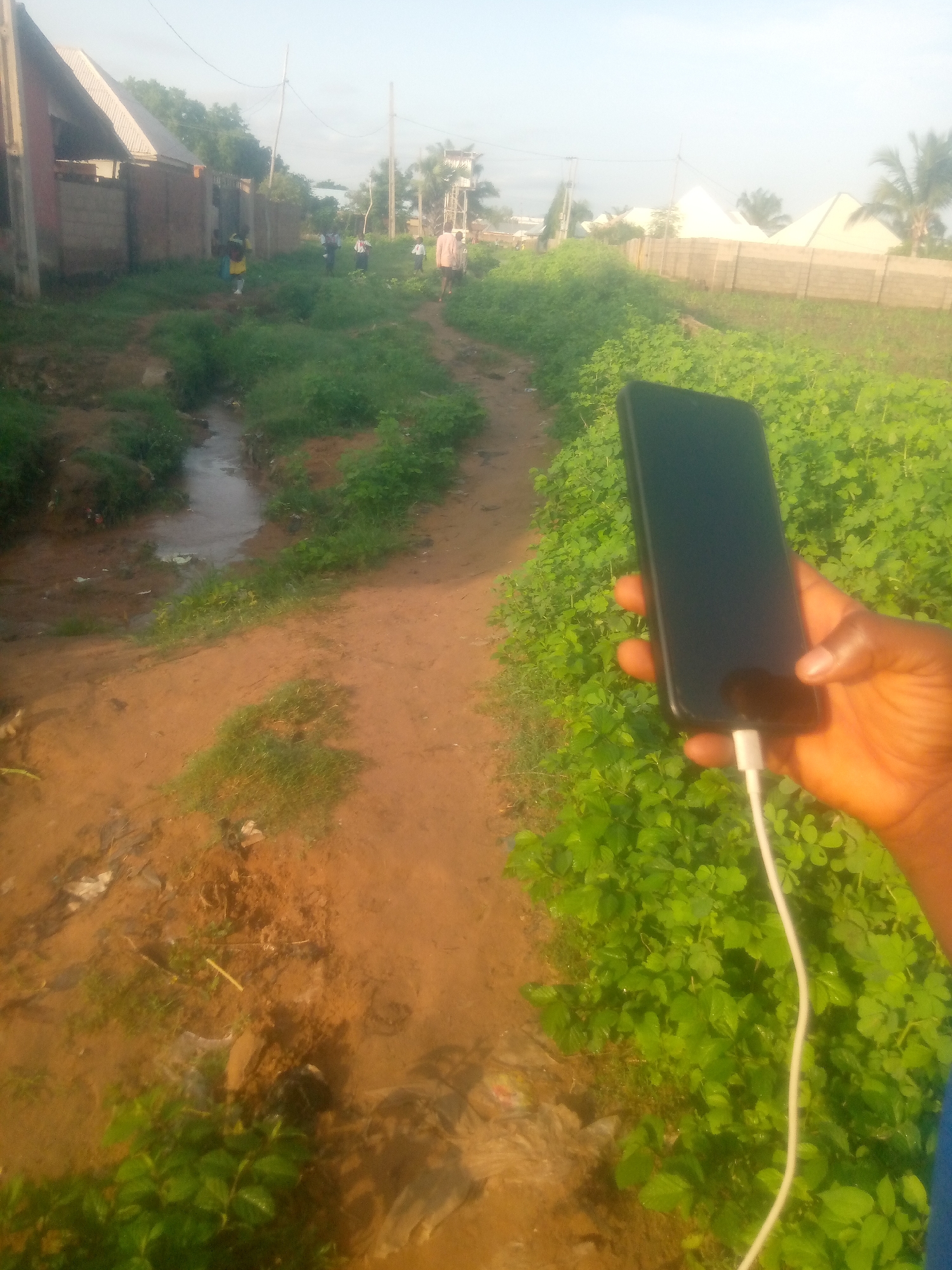 A first-person perspective shot shows a hand holding a black smartphone with a white charging cable plugged into it. The hand is dark-skinned and is positioned in the lower right quadrant of the image. The phone screen is dark and reflects some of the surrounding green foliage. The scene appears to be outdoors in Jauro Yinu, Nigeria. A dirt path curves through an area with abundant green vegetation on the right and a small, muddy stream on the left. In the background, a few people, including children in yellow and blue clothing, can be seen walking along the path. Buildings with corrugated metal roofs and brick walls are visible on the left side of the path. The sky is bright and appears to be overcast or hazy.
