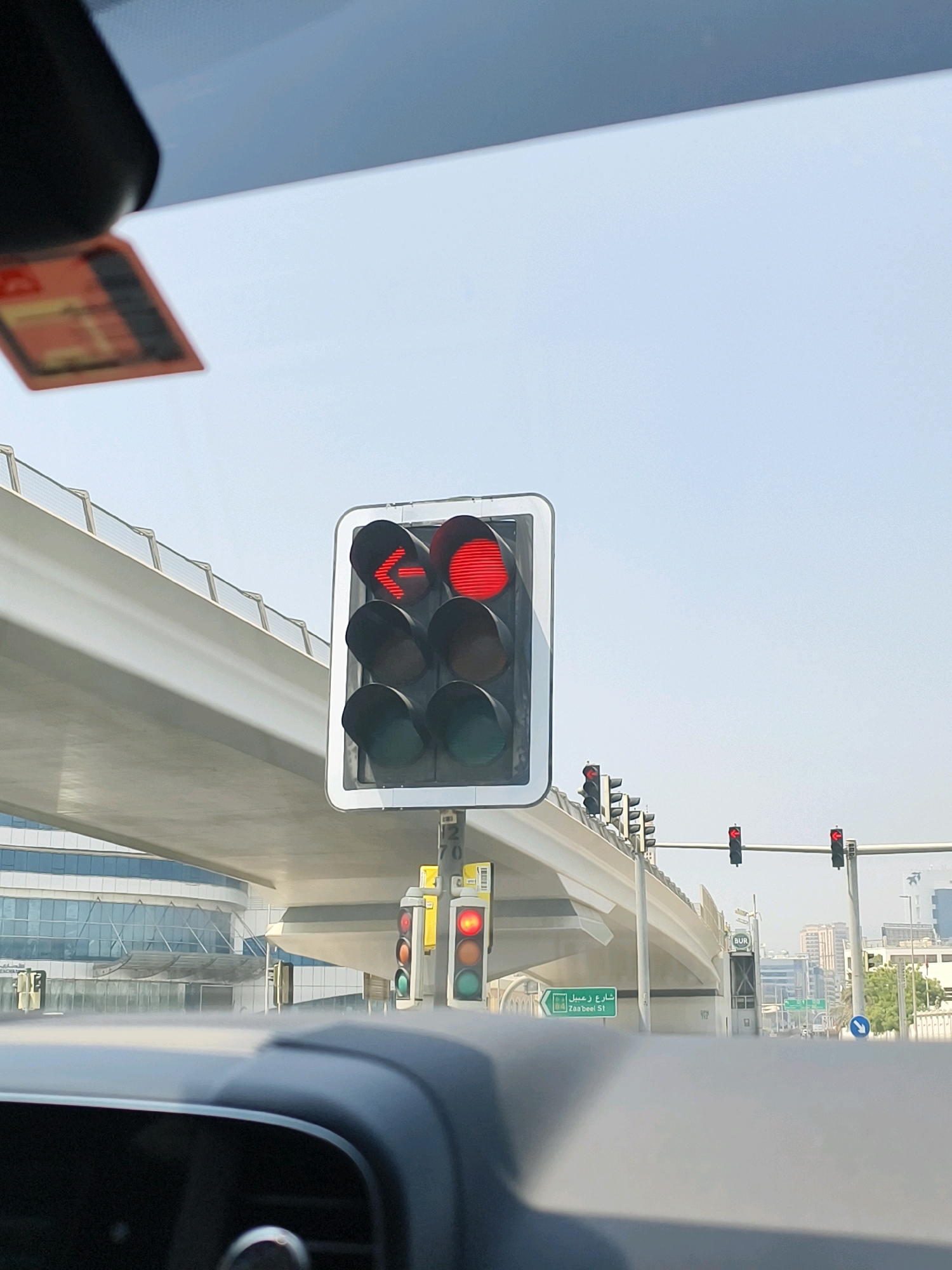 The image captures a street-level view from within a vehicle, looking towards a traffic intersection in Dubai, United Arab Emirates. The prominent subject is a traffic light displaying a red left-turn arrow and a solid red circle, indicating that vehicles must stop and that left turns are prohibited. Additional traffic lights are visible further down the road, also showing red signals. The environment features modern urban architecture, including an elevated highway structure and contemporary buildings with glass facades. A street sign in Arabic and English, indicating "Zaa'beel St," is also visible. The sky is clear and bright, suggesting a daytime scene. The image is framed from the perspective of a driver or passenger, with the car's dashboard and windshield partially visible in the foreground.