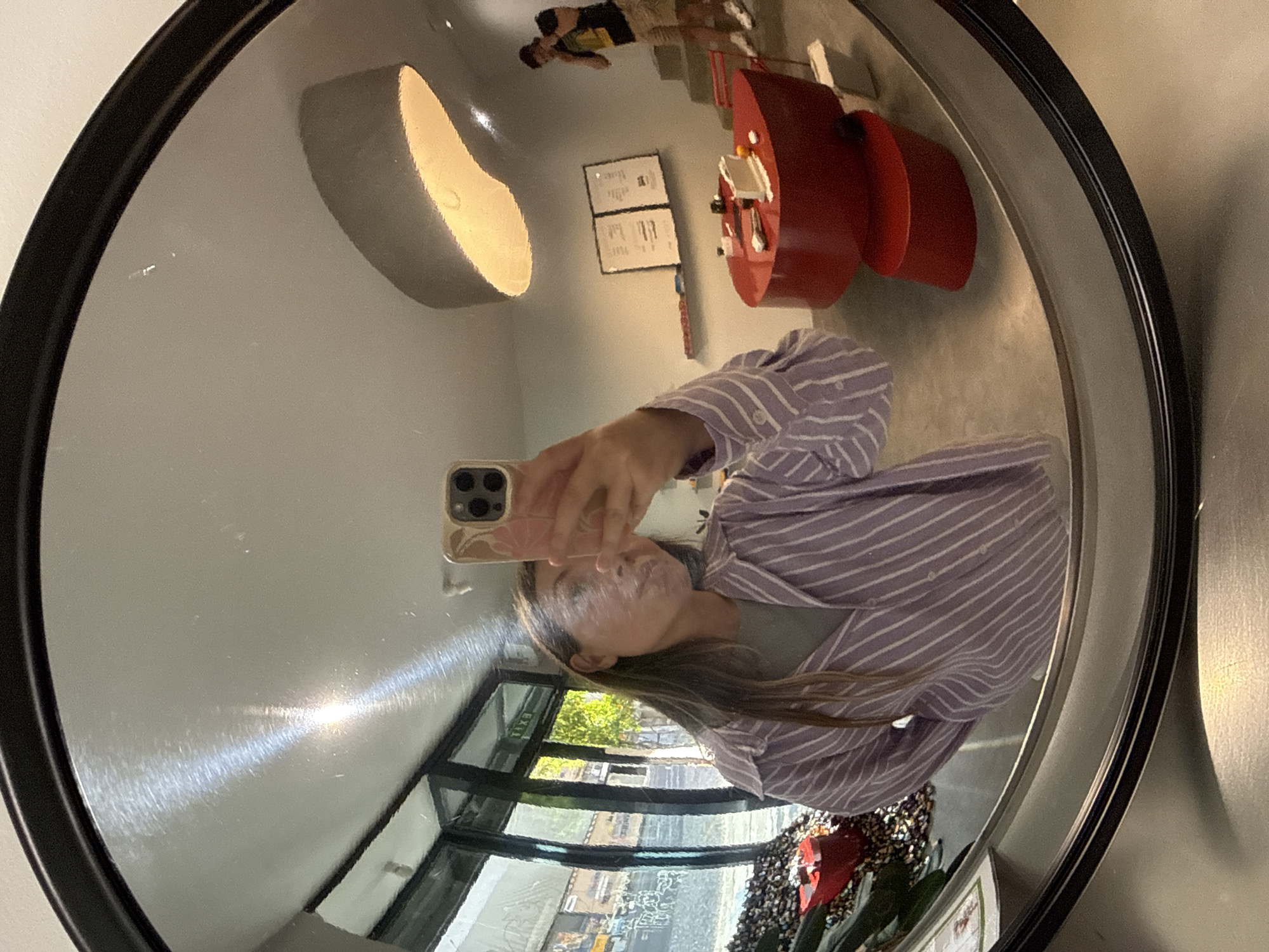 The image shows a reflection in a round mirror with a black frame. The reflection captures a person taking a selfie with a smartphone. The person is wearing a striped shirt. The background includes a window with a view of a city street, indoor plants, and other reflections of interior elements, including furniture, a light fixture, artwork on the wall, and another person in the distance. Given the presence of Cyrillic characters visible through the window, it is highly probable that the location is in the city of Belgrade, Serbia.