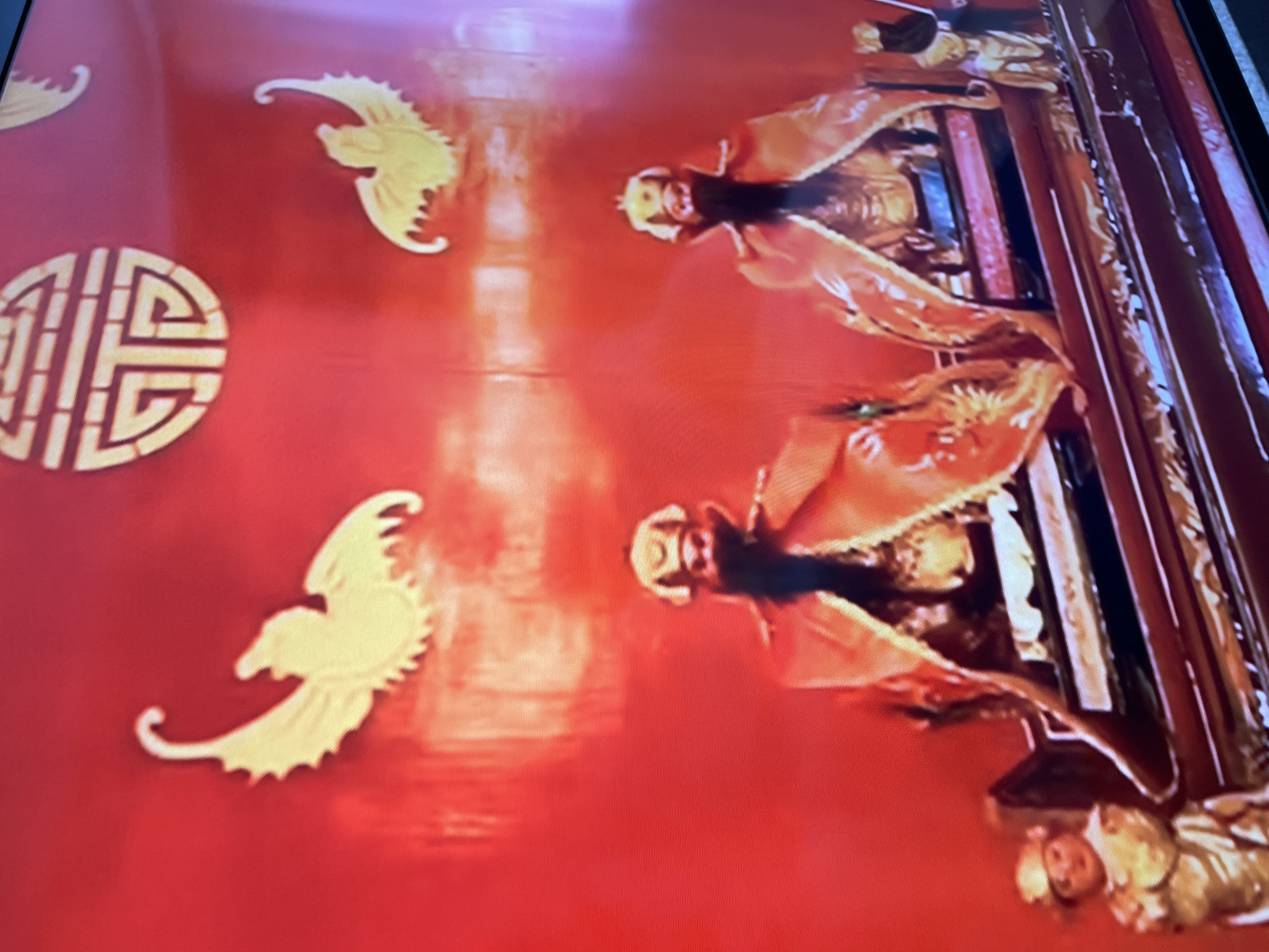 The media file captures a vibrant, richly decorated indoor scene, likely a cultural display or installation, set in Moscow, Russia, with a distinct Chinese aesthetic. The dominant color is a striking red, forming the background, possibly a wall or large screen.
The primary subjects are two prominent figures, appearing to be statues or mannequins, positioned on the right side of the frame. They are depicted in elaborate, traditional Chinese attire, featuring voluminous robes in shades of orange and gold, with intricate patterns and details. Both figures wear ornate golden headwear and have dark, long hair or beards, conveying a sense of majesty or historical significance. Their expressions are neutral. A smaller, less distinct figure, also in traditional dress, is visible at the very bottom right, part of the same display structure.
The red background is adorned with several golden, stylized phoenix-like birds with flowing tails, adding to the opulent and auspicious theme. Prominently featured on the left side is a circular golden emblem containing the Chinese character "福" (Fú), which symbolizes "fortune" or "good luck."
The scene appears to be a static exhibit, with no active event taking place. The indoor setting precludes any discernible time of day or weather conditions. The overall ambiance is grand and culturally specific, suggesting a celebration or presentation of Chinese heritage within a Russian context. The image quality indicates it might be a photograph of a screen or a brightly lit projection, given the visible pixelation and reflection.