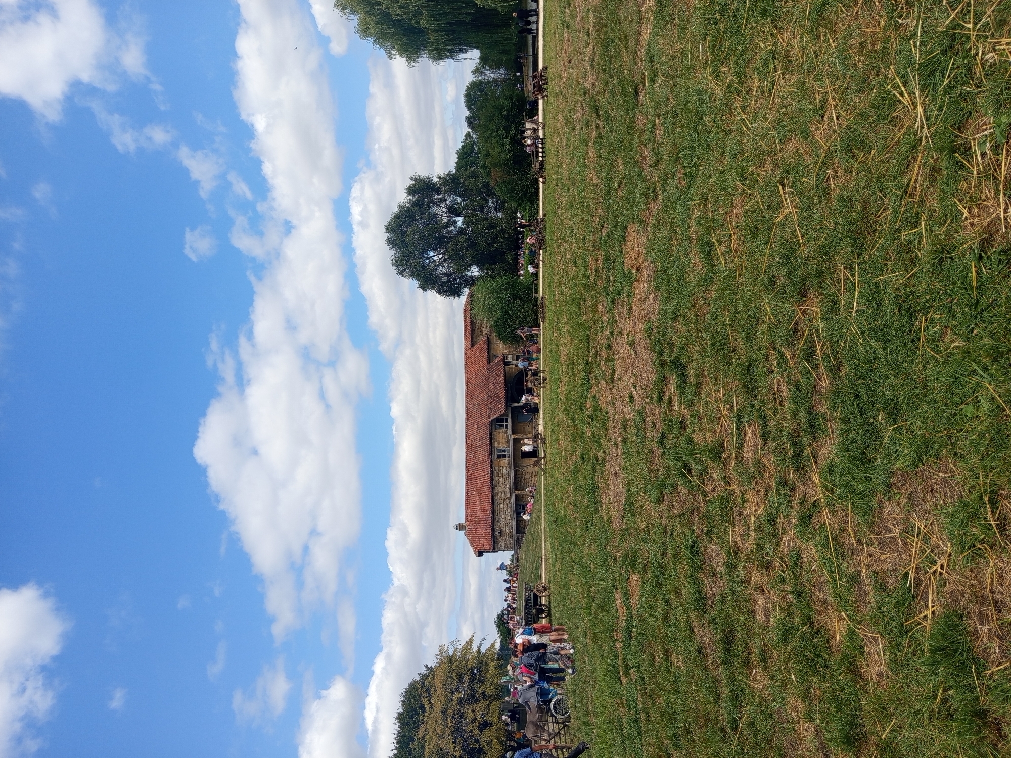 The image depicts an outdoor scene at a historical reenactment or museum in Azannes-et-Soumazannes, France. A large, traditional building with a red tile roof and stone walls is visible in the background, situated on a gentle slope. Several people, including adults and children, are gathered around the building and in the surrounding grassy area. Some are observing exhibits or demonstrations, while others appear to be casually interacting. In the foreground, a mowed grassy field with patches of dry straw is present. Various old farm implements and carts are scattered across the landscape, suggesting a historical farming context. The sky is a bright blue with scattered white clouds. The overall impression is of a sunny day at a public event focused on historical preservation and education.