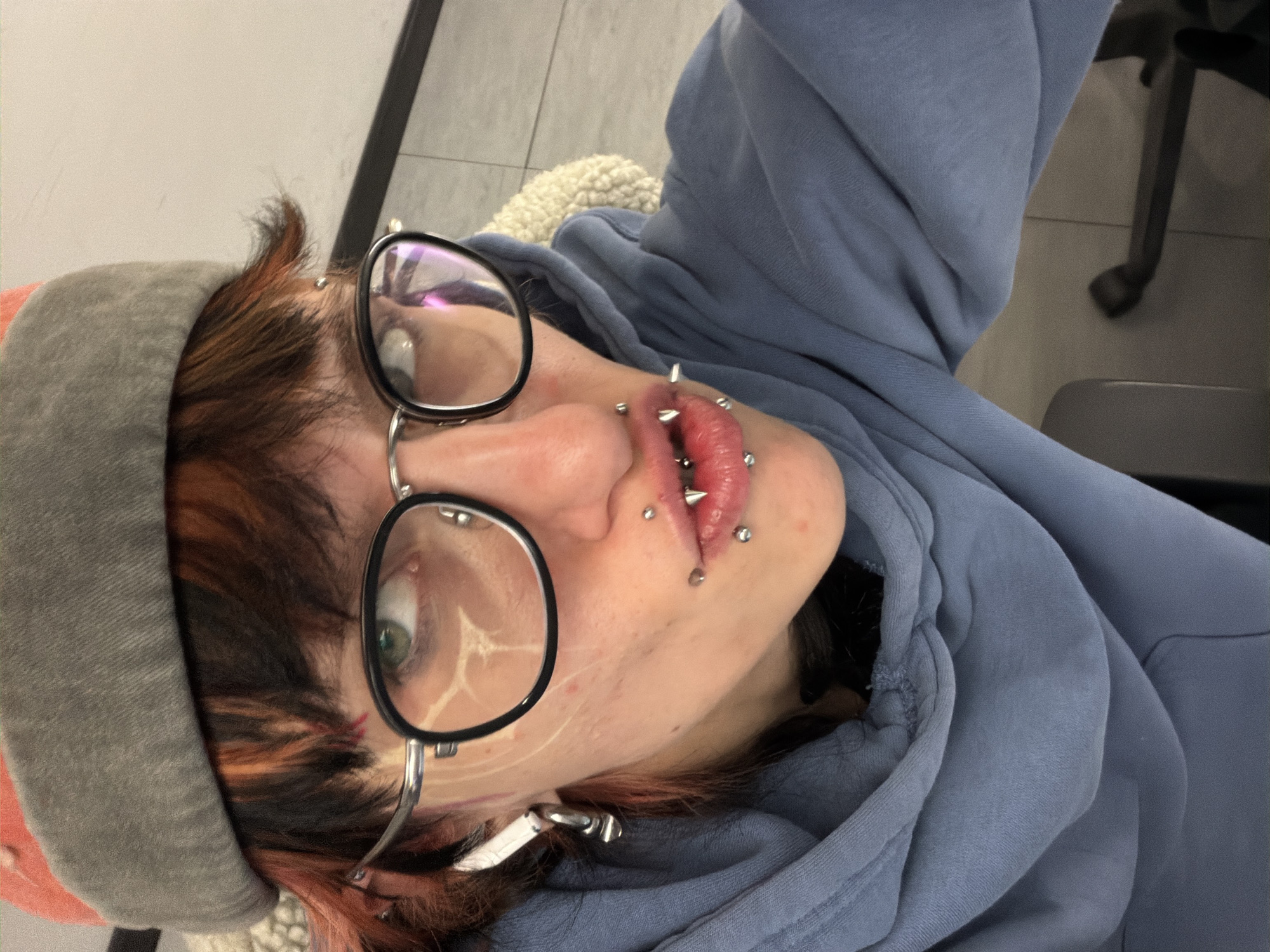 A close-up, tilted selfie captures a person with striking facial modifications. The individual wears round, black-rimmed glasses and a gray beanie. Their hair is multi-toned, with shades of brown, black, and red/orange highlights. Numerous piercings adorn the lips, including spike-shaped jewelry. The person is wearing a blue hoodie. The background shows glimpses of an interior space with neutral-colored flooring and what appears to be office furniture. The location is not discernible as Sofia, Bulgaria, without further clues.
