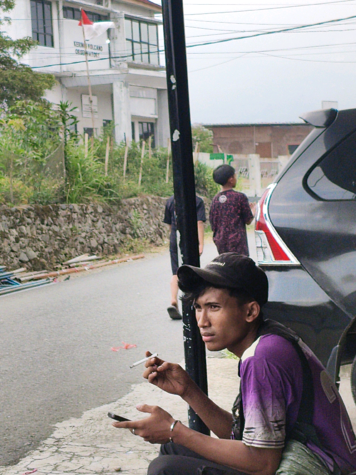 In Lindung Jaya, Indonesia, a young man wearing a black cap and a purple t-shirt sits on the side of a road, holding a cigarette in his right hand and a mobile phone in his left. He is looking to his left, away from the camera. The road is paved and appears to be in a semi-urban or rural area. Behind him, a dark-colored car is partially visible. In the background, there is a building with a white facade and dark windows, and dense green vegetation. A stone wall lines the side of the road. A thin, dark pole is positioned in the foreground, partially obscuring the view of the young man and the car. The sky is overcast.