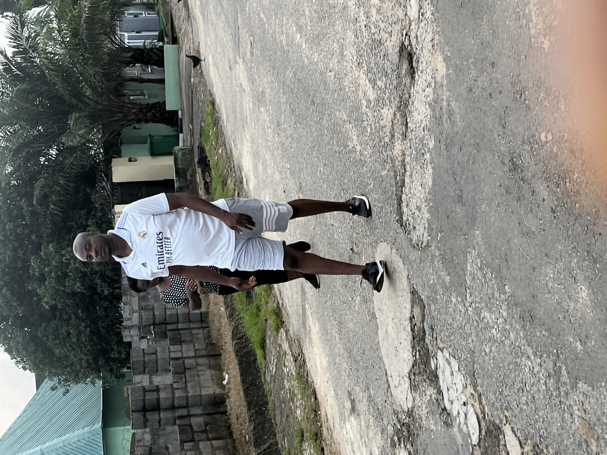A man wearing a white short-sleeved shirt with an "Emirates Fly Better" logo and grey shorts walks on a paved road in Abuja, Nigeria. He is also wearing black and white sneakers. Behind him, a woman in a black and white polka dot shirt walks. In the background, there are buildings, palm trees, a green trash can, a pile of grey bricks, and two chickens on the road. The road has several potholes and cracks. The sky is overcast.