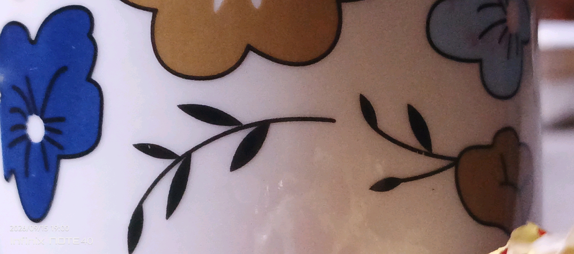 The image displays a close-up view of a white ceramic mug decorated with colorful floral patterns and black vine-like branches with leaves. On the left side, a blue flower with black outlines and black lines radiating from its center is visible. To the right of the blue flower, black branches with oval-shaped leaves extend across the mug. Further to the right, there are portions of brown and light blue floral designs. The background is a pale, textured surface, likely the mug itself, with subtle lighting. In the bottom left corner of the image, a timestamp and device information are overlaid: "2026/09/15 19:00" and "Infinix NOTE 40". The location in Muyan, Uzbekistan, is not discernible from the visual content of the image.