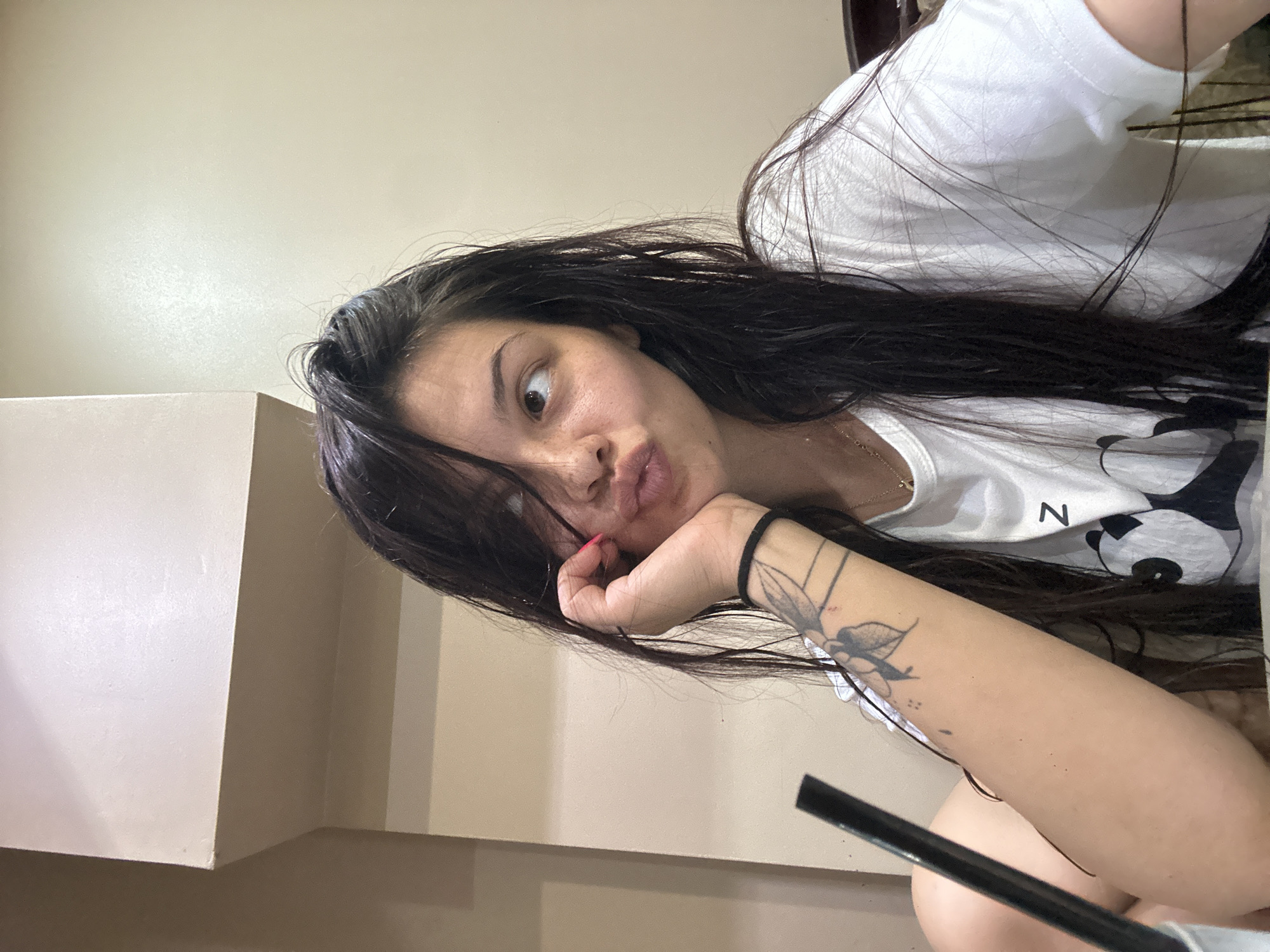 The media file is a selfie of a young woman with long, dark hair. She is wearing a white t-shirt with a panda graphic. The woman is resting her chin on her hand, and her eyes are wide with surprise. Her lips are slightly pursed. She has a tattoo on her left forearm that appears to be of a flower. A thin gold necklace is visible around her neck. The background is a plain, light-colored wall, suggesting an indoor setting. The lighting appears to be natural, possibly from a window, as there are some shadows and highlights on her face. The image is oriented sideways. There is a small black object, possibly a straw or pen, in the lower right corner of the frame. The overall mood of the image is one of playful surprise or curiosity. No specific time of day or weather conditions are discernible. No text is clearly legible beyond the graphic on her shirt. The provided location context of Sliven, Bulgaria does not offer any specific visual cues within the image itself.