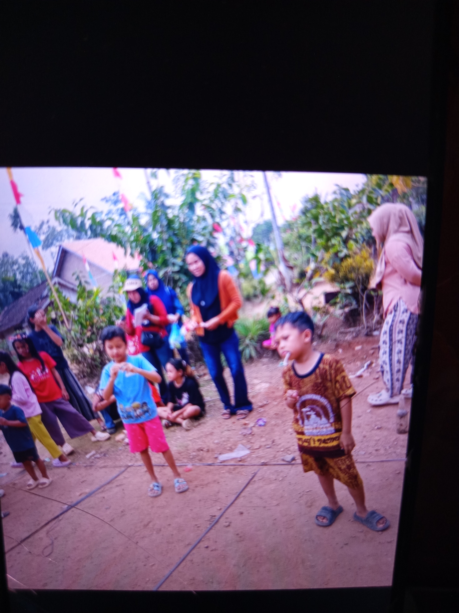 The image depicts an outdoor gathering in a rural area, likely in Indonesia, with several people, mostly children, present. In the foreground, a young boy in a yellow and brown patterned t-shirt and matching shorts is standing, holding something to his mouth. To his left, another child in a blue t-shirt and pink shorts is also standing. Further back, a group of women and children are visible, some wearing headscarves. One woman in a blue headscarf and orange cardigan is standing while holding a small bag. Another woman in a tan headscarf and patterned pants is standing further to the right. The ground appears to be dirt, with some haphazardly laid out black ropes or wires creating a diamond shape on the ground. In the background, there are trees, foliage, and a simple structure that could be a house. Colorful flags or banners are strung across the area, suggesting a festive occasion or event. The overall scene appears informal and casual, possibly a local community event or celebration.