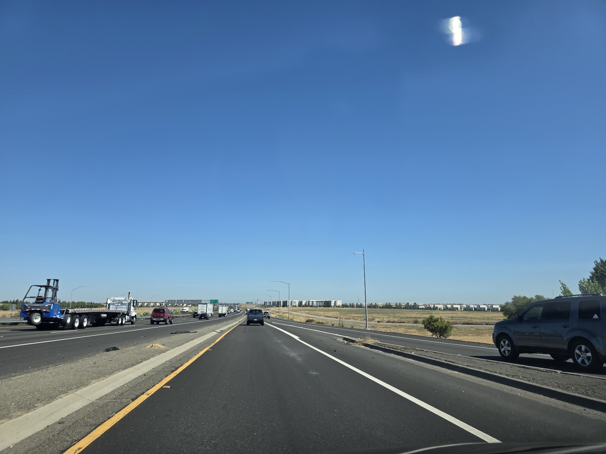 The image depicts a clear, sunny day with a deep blue sky. The perspective is from inside a vehicle traveling on a highway, likely in or near Sacramento, California, based on the vegetation and urban sprawl visible in the distance.
The highway has multiple lanes and is paved with asphalt. White and yellow lane markers are visible. To the left, a large blue forklift is being transported on a flatbed truck. Other vehicles, including cars and trucks, are also on the highway.
The environment includes dry, grassy fields on either side of the highway, with a line of trees and distant buildings that suggest a suburban or exurban area. A streetlamp stands tall on the right side of the road. The overall scene is one of ordinary highway travel on a bright day.