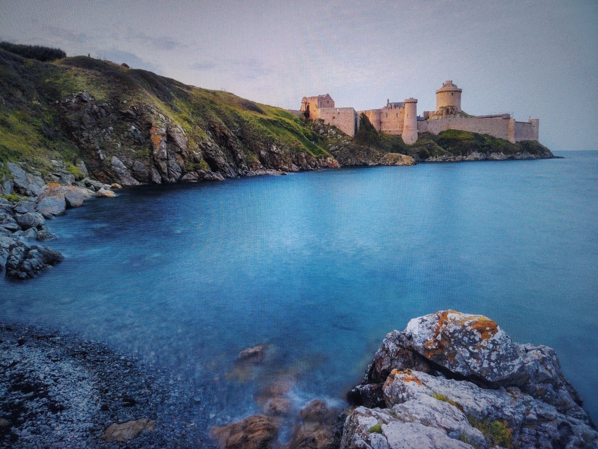 The image depicts a vast expanse of deep blue ocean meeting a rugged, rocky coastline. On the cliff edge overlooking the sea, a formidable stone castle stands prominently. The castle features multiple towers, including a central circular one, and is constructed from weathered, light-colored stone. The coastline is characterized by steep, grassy slopes interspersed with rocky outcrops and a small, dark pebble beach in the foreground. The water appears calm, with subtle ripples and some blurred rocks visible beneath the surface near the shore. The sky above is overcast with soft, diffused light, contributing to a serene, yet dramatic atmosphere. The location of this scene is not in Tokmok City, Kyrgyzstan.