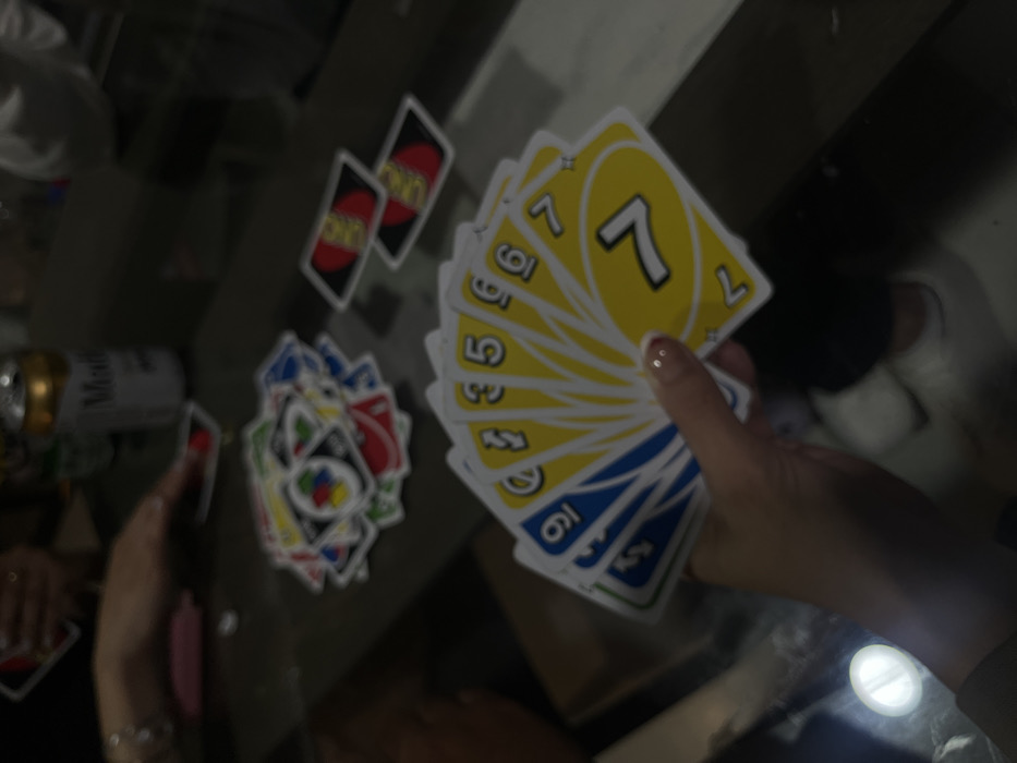 A slightly blurred image shows a hand holding several Uno cards, the top one being a yellow 7. A pile of discarded Uno cards sits on the table. Two red "Uno" cards are also visible nearby. A can of Modelo beer is on the left side of the table. The image appears to be taken from above, showing hands and a table surface.