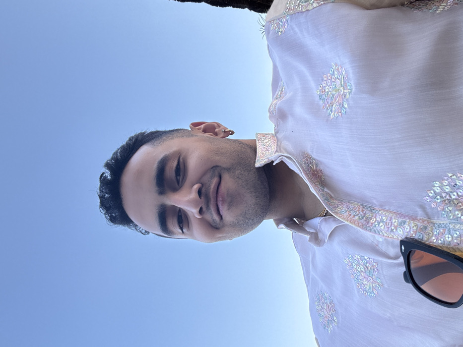 A close-up, low-angle portrait features a male individual positioned centrally, looking directly at the camera with a slight smile, against a clear light blue sky. The individual, appearing to be of South Asian descent, wears a light pink or off-white kurta adorned with iridescent sequin and embroidered patterns along the band collar, placket, and in floral motifs on the chest. A gold-colored chain is visible around his neck, and a small gold-colored hoop earring is present in his left ear. Black sunglasses with orange-tinted lenses are hooked onto the right side of his kurta's placket. His hair is dark, short, and curly, and he has short facial hair. A dark, out-of-focus vertical element is partially visible on the far right background. The scene is located in the city of Malasar, India.