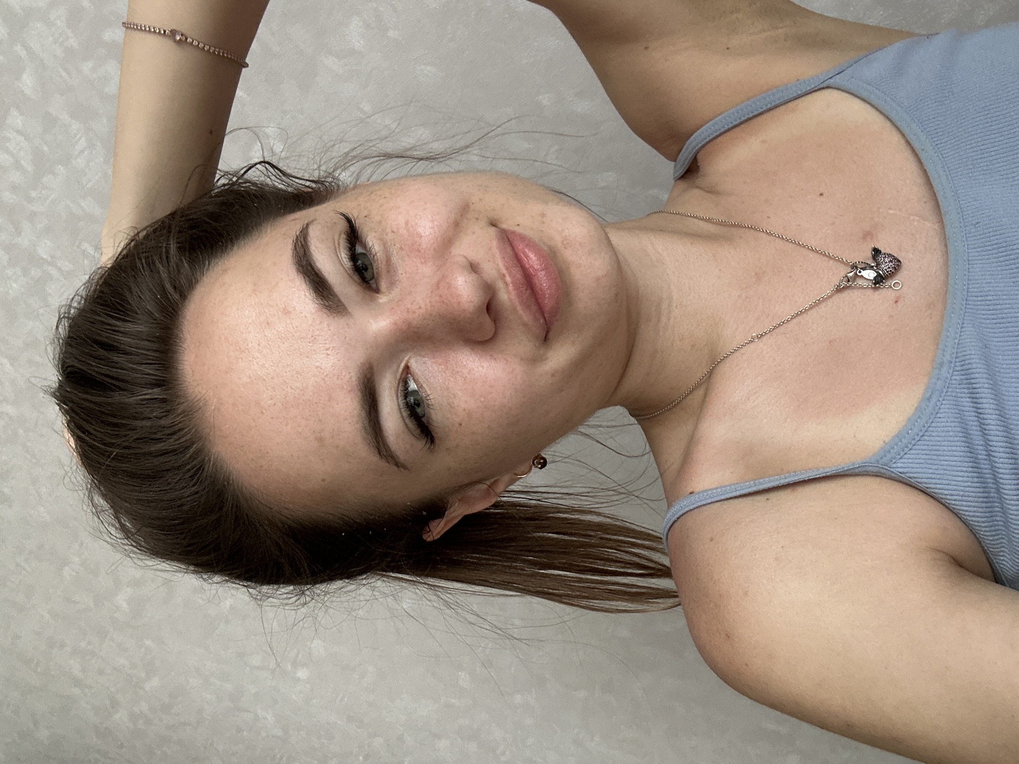 An indoor, medium shot shows a young Caucasian woman with brown hair pulled back in a ponytail, her right hand touching the back of her head. She has green eyes, freckles across her nose and cheeks, and is wearing a light blue tank top with thin straps. A silver chain with a charm pendant is around her neck. A beaded bracelet with a small pink heart charm is on her wrist. The background is a textured gray wall.