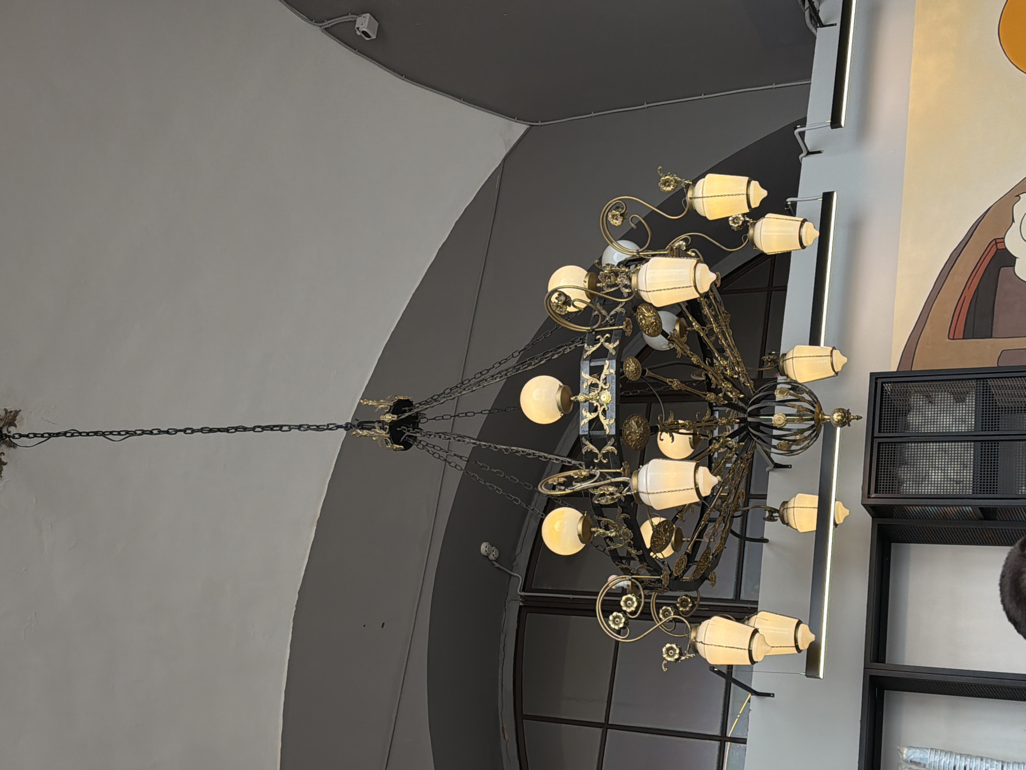 The image shows an ornate chandelier hanging in what appears to be a public space. The chandelier is made of dark metal with brass-colored accents and features multiple white cylindrical light fixtures. The architecture suggests a modern building with a curved ceiling and large windows. The location is Moscow, Russia. The image is taken from below, looking up at the ceiling and chandelier. The environment looks like a commercial or public building in an urban area.