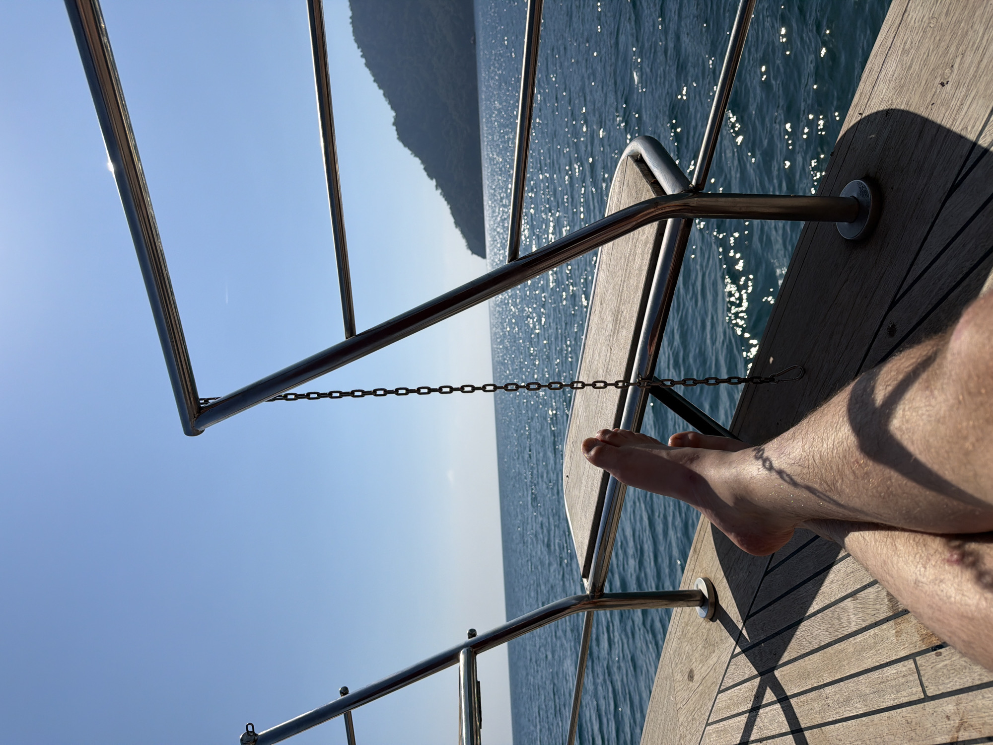 The image captures a bright, sunny day at sea from the perspective of someone relaxing on a boat in Kumluca, Turkey.
In the foreground, the bare lower legs and feet of a person are stretched out over a light brown wooden boat deck, suggesting a relaxed posture. The skin appears tanned, and there are strong, dark shadows cast by the legs and chrome railings onto the deck. Subtle glitter or sparkling specks are visible on one of the legs.
The boat itself features a teak deck with distinct wooden planks and shiny chrome or stainless steel railings that line the vessel's edge. A metal chain is visible, hanging between two sections of the railing.
Beyond the boat, the deep blue sea shimmers brightly with numerous sparkling reflections from the strong sunlight. In the distant background, a dark green mountain or landmass rises from the water, silhouetted against a clear, light blue sky. The sky also shows a faint sun glare and a wispy contrail.
The activity depicted is one of leisure and relaxation, with the person enjoying the sunny weather and scenic maritime view. The strong sunlight and clear sky indicate daytime, likely mid-day or afternoon, with fair weather conditions. There is no visible text in the image.