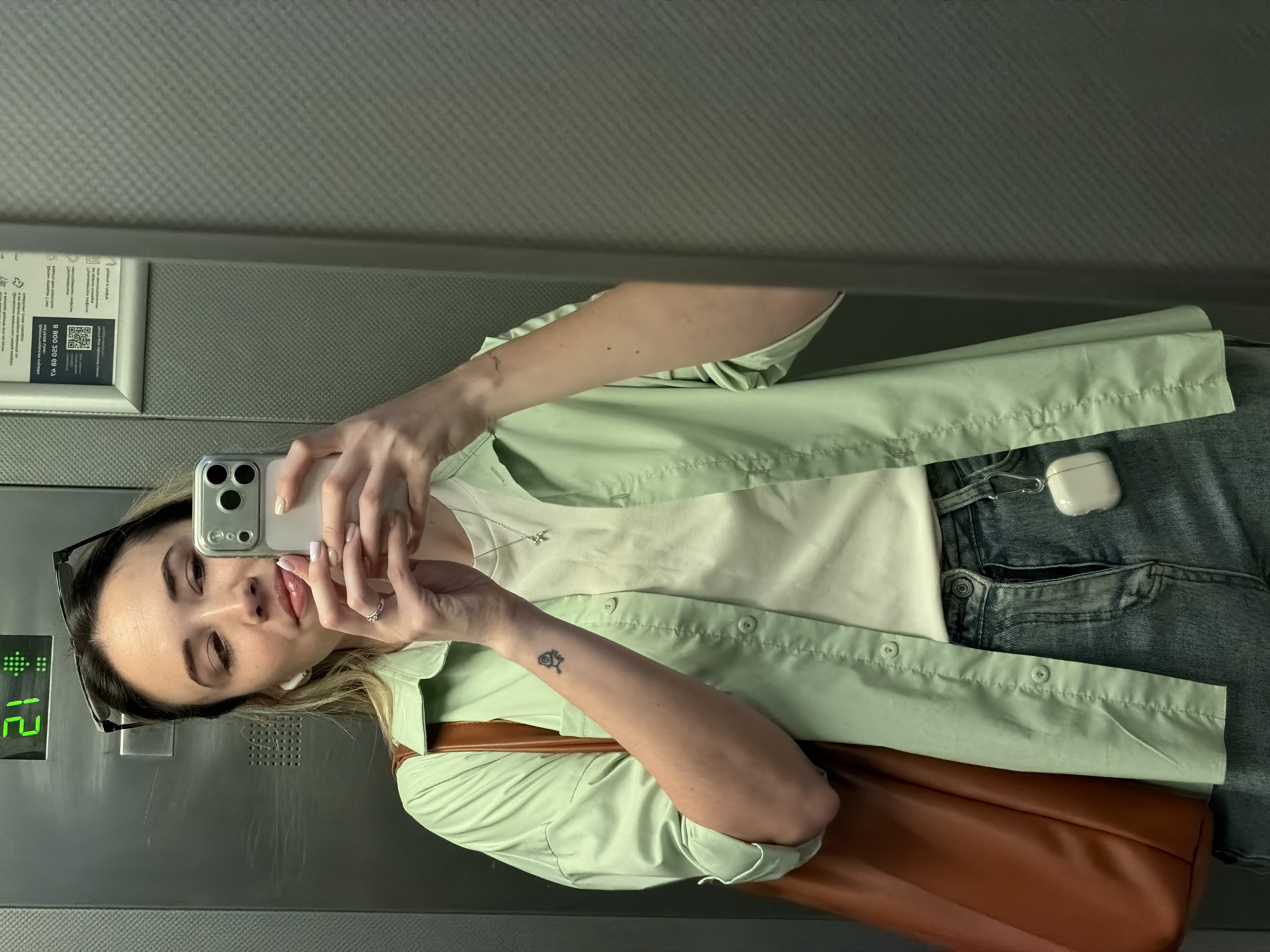 A young woman is taking a selfie in an elevator, possibly in Kaliningrad, Russia, though specific visual confirmation of the city is absent. She holds an iPhone in her right hand, her left hand also visible near the phone. She is fair-skinned with light brown hair, wearing a white t-shirt, a light green button-down shirt, and blue jeans. A brown shoulder bag rests on her shoulder. Sunglasses are perched on her head. An AirPod case is visible on her jeans. The elevator's digital display shows the number "12". A sign with a QR code is visible on the elevator wall. The elevator's interior is paneled in a neutral color. A small tattoo is visible on her left forearm. She wears a ring on her left ring finger.