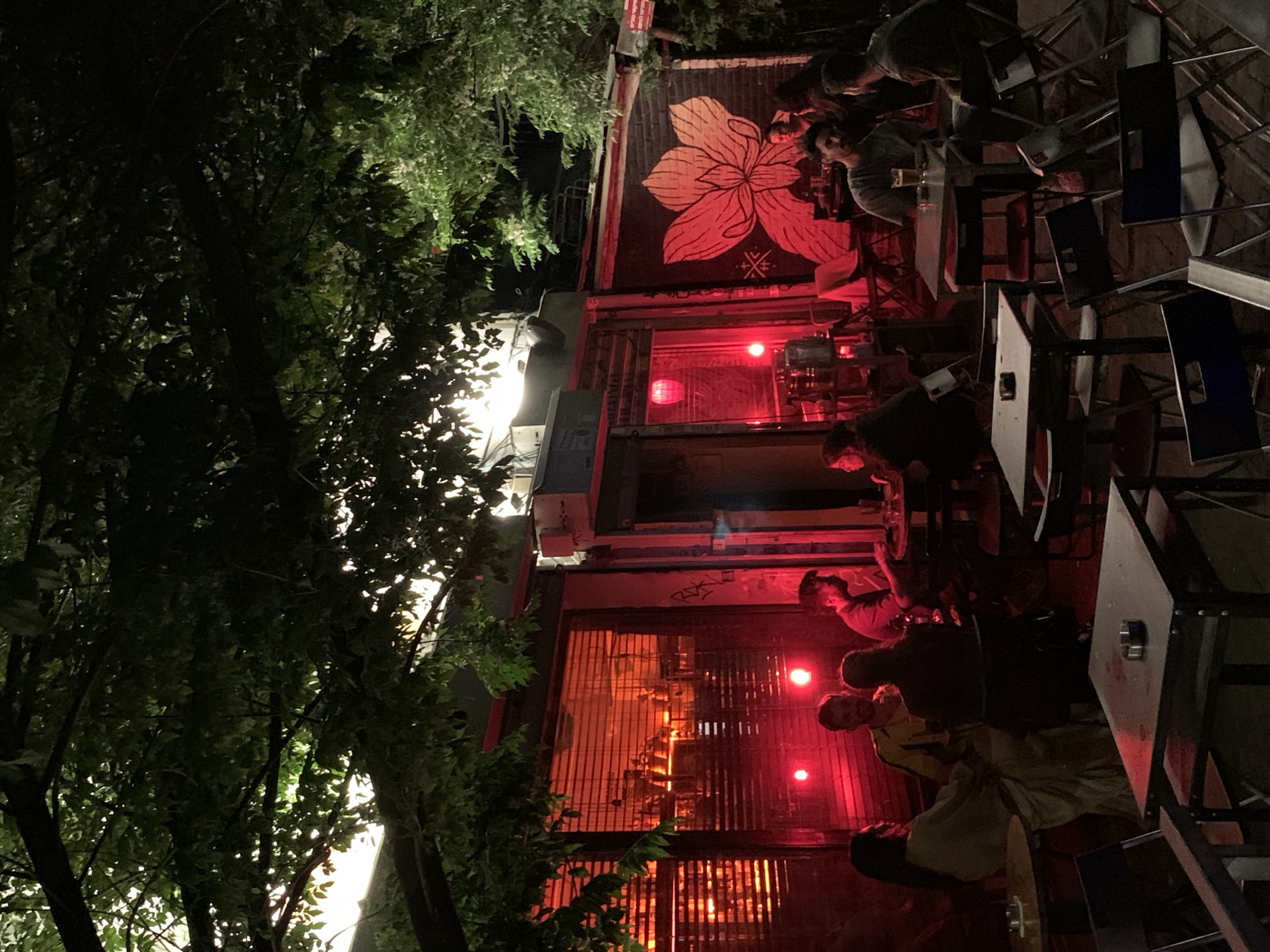 At night, an outdoor bar in Tel Aviv, Israel, shows people seated at metal tables and chairs. The bar features red lighting and a mural of a flower. Green trees are visible on the left side of the frame. The scene appears to be a gathering of people enjoying the nightlife.