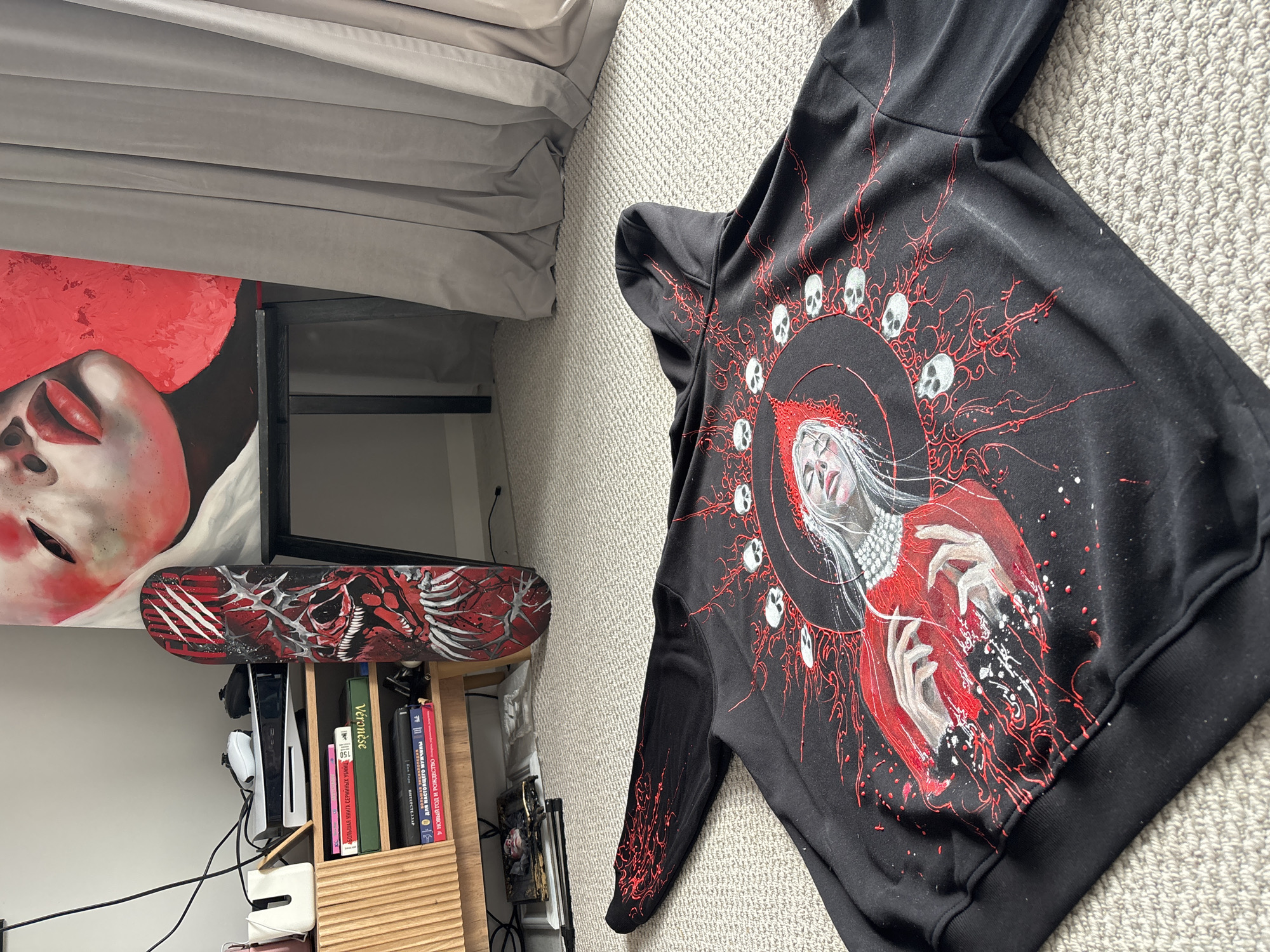 The image displays an indoor scene, likely a personal room in Krasnoyarsk, Russia, filled with various objects. In the foreground, a black hooded sweatshirt with a detailed, gothic-style graphic print is spread out on a light grey carpet. The print features a central figure, possibly a woman or saint, with white hair, a red garment, and pale hands, encircled by a halo of white skulls. Red, intricate, vine-like patterns and white splatters radiate from this central image, extending onto the sleeves.
To the left, a grey upholstered bed or sofa is partially visible. Against the wall, above a wooden bookshelf, a painting of a woman's face is displayed. She is depicted with bold red lips, pale skin, and a large red hat or headpiece. Below the painting, a black skateboard rests horizontally, showcasing a graphic of a snarling red, black, and white creature with prominent claws and skeletal features.
The wooden bookshelf holds a white PlayStation 5 console, two white controllers, and several books. Visible titles on the books include "АБОЛЬШАЯ КНИГА СЕРИЙНЫХ УБИЙЦ" (A Big Book of Serial Killers), "О ДЛЯ НАСТОЯЩЕГ НАСТОЯЩЕГО МУЖЧИНЫ" (For the Real Man), "У НОВЫЙ ГОД И РОЖДЕСТВО" (New Year and Christmas), and "Véronèse". The room's contents suggest interests in gaming, art, and possibly dark or alternative culture. No specific activity is depicted; the items are stationary, displayed within the room. The lighting appears to be ambient, indicative of daytime or a well-lit interior.