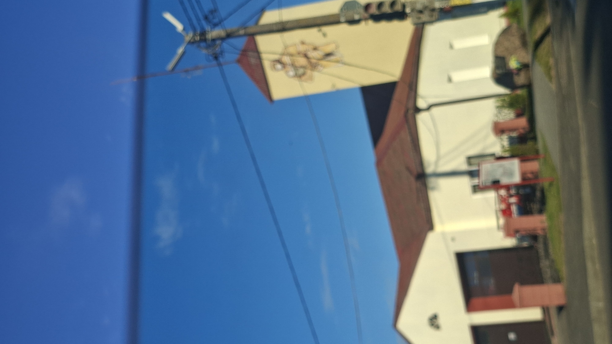 The image, taken on a clear day, shows a building with a red roof and white walls. A tall, yellow tower with a red roof is attached to the building, and features a depiction of two figures. A utility pole with wires stands to the right of the tower. In front of the building, there are several red pillars and a sign. A dark-colored vehicle is visible in one of the garage bays of the building. The scene is located in the city of Smaliavicy, Belarus.
