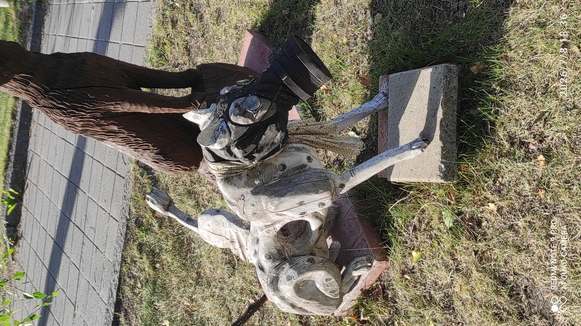 The image shows a wooden sculpture of a dog wearing a gas mask and a bandana, placed outdoors on a grassy area with some scattered leaves. Behind the dog sculpture, a portion of another dark wooden sculpture of a sitting animal, possibly a bear or a larger dog, is visible. The ground is covered with dry grass and some fallen leaves. In the background, a paved walkway with square tiles and a shadow cast by sunlight can be seen. The overall lighting suggests it is daytime. The image contains a watermark at the bottom left corner reading "REDMI NOTE 9 PRO AI QUAD CAMERA" and a timestamp at the bottom right corner reading "2026/7/8 18:36". The location in Iisalmi, Finland is not specified in the image itself, but this is a general description of the visual content.