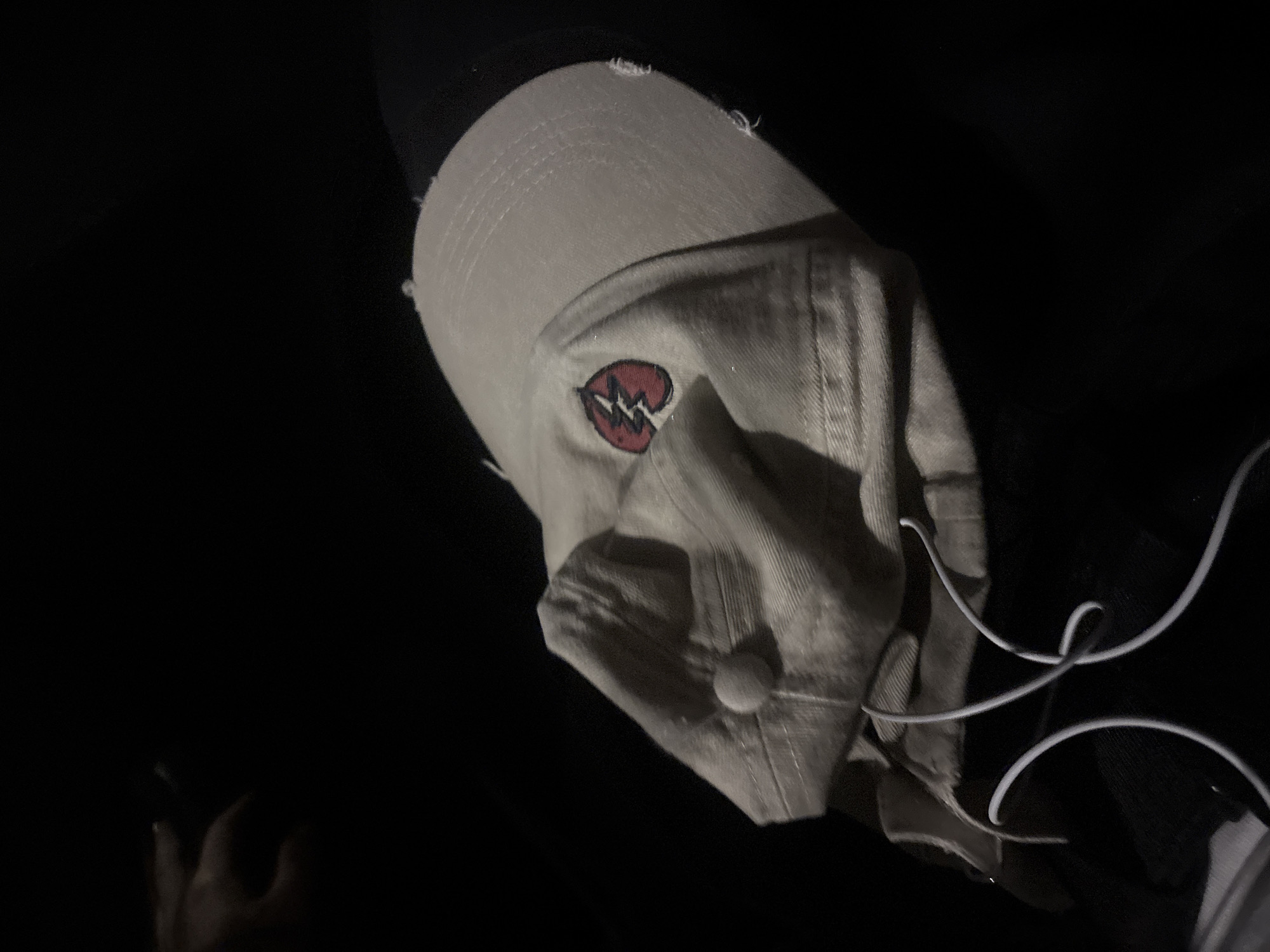 A beige baseball cap with a red broken heart design is resting on a dark surface. Part of a black object is visible behind the cap, and white earbuds are tangled to the right. A finger is slightly visible at the bottom left corner of the frame. The location is unknown, but a city has not been indicated.