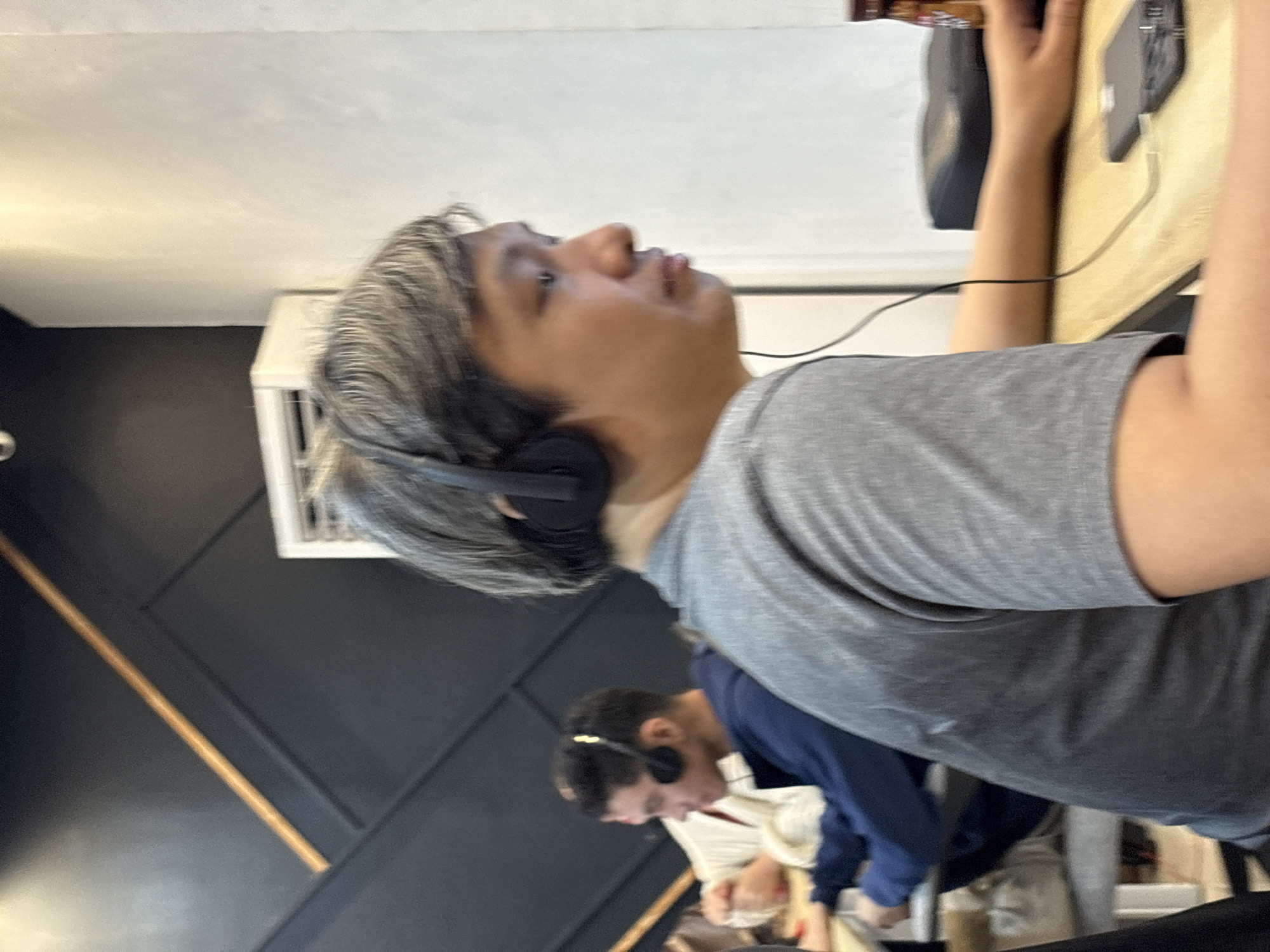 In a coffee shop in Davao City, Philippines, two individuals are seated at a table. The person in the foreground, facing right, wears a grey t-shirt and headphones, with a phone and a bottle visible on the table. The second person, in the background, facing left, wears a blue sweatshirt and headphones, with a white object and their hands on the table. The shop features dark walls with geometric wooden accents and a light-colored wall on the right.