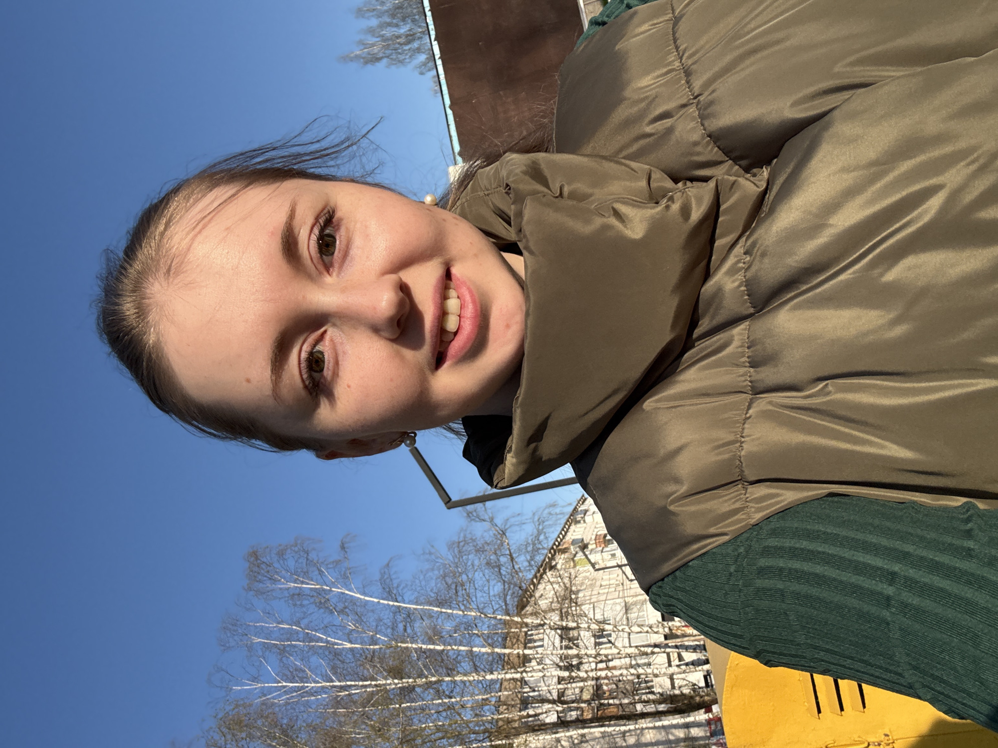 A young woman is taking a selfie outdoors. She is wearing a brown puffy jacket and a dark green ribbed shirt. She has light brown hair pulled back from her face, and she is smiling. In the background is a clear blue sky, bare trees, and a building with a white facade and multiple windows. A yellow object is visible in the lower right corner of the frame.