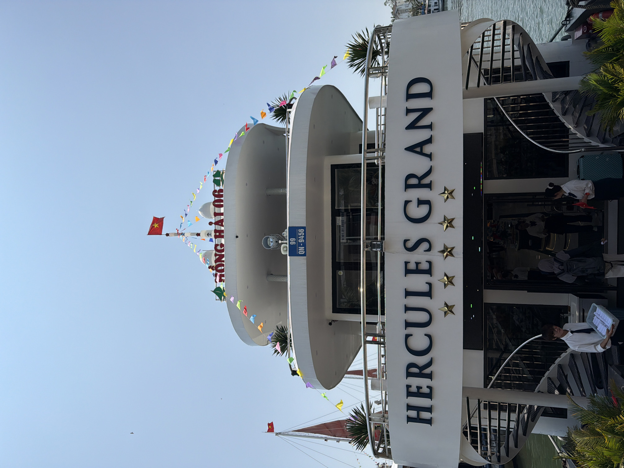The image shows a vertical view of the Hercules Grand boat in Tuan Chau Ward, Vietnam. The boat has a multi-tiered white structure with colorful flags strung around its edges. "HERCULES GRAND" is printed vertically on a white surface with three gold stars. People can be seen near the entrance, including a person in a white shirt and black tie carrying a basket. A Vietnamese flag flies atop the boat. A sign displaying the registration number "99 QN-9456" is visible. In the background, the sky is clear blue.