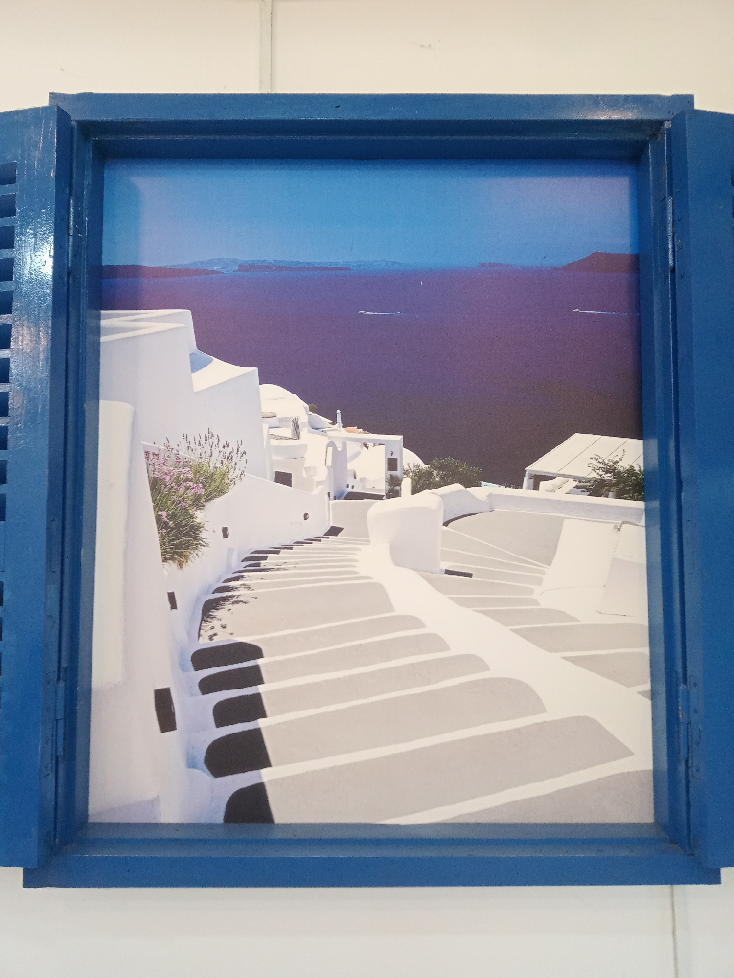 The image shows a framed print of a typical Santorini landscape with whitewashed buildings and stairs leading towards the sea. The frame is blue and appears to mimic a window shutter, which gives the impression of looking at the view through a window. There are no people visible in the image. The environment is Santorini with white stairs and buildings on the island. The location of the photograph is likely in an interior setting, given the white wall behind the framed picture. The information regarding Nha Trang, Vietnam, is irrelevant as the image clearly depicts Santorini.
