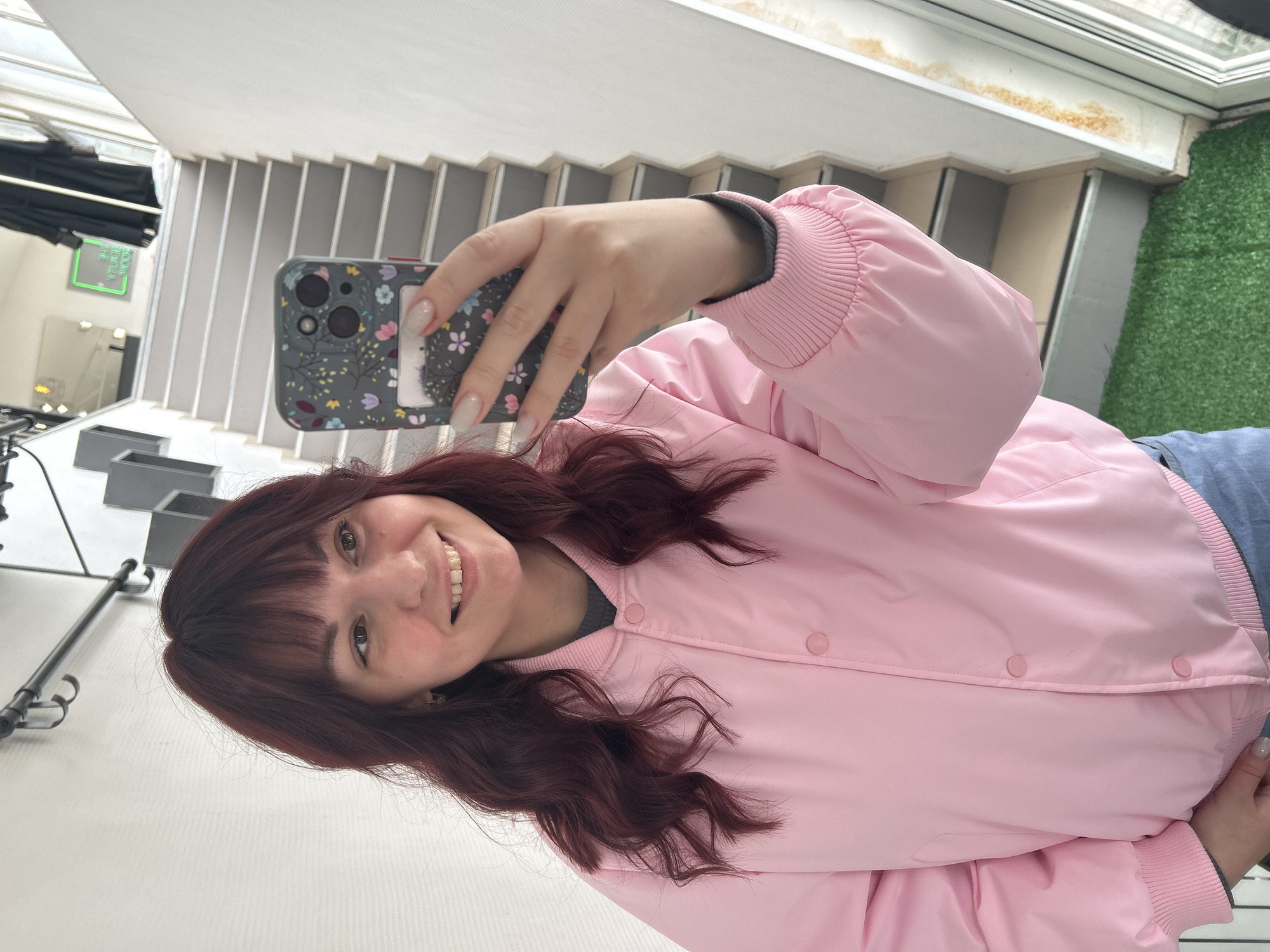 A young woman is taking a selfie indoors. She has auburn hair with bangs, and is wearing a pink bomber jacket. She is holding a phone with a floral case up in her right hand, with a staircase in the background. Also visible is a window with green turf visible on the other side and lighting equipment. The image was taken in Chaikovsky, Russia.
