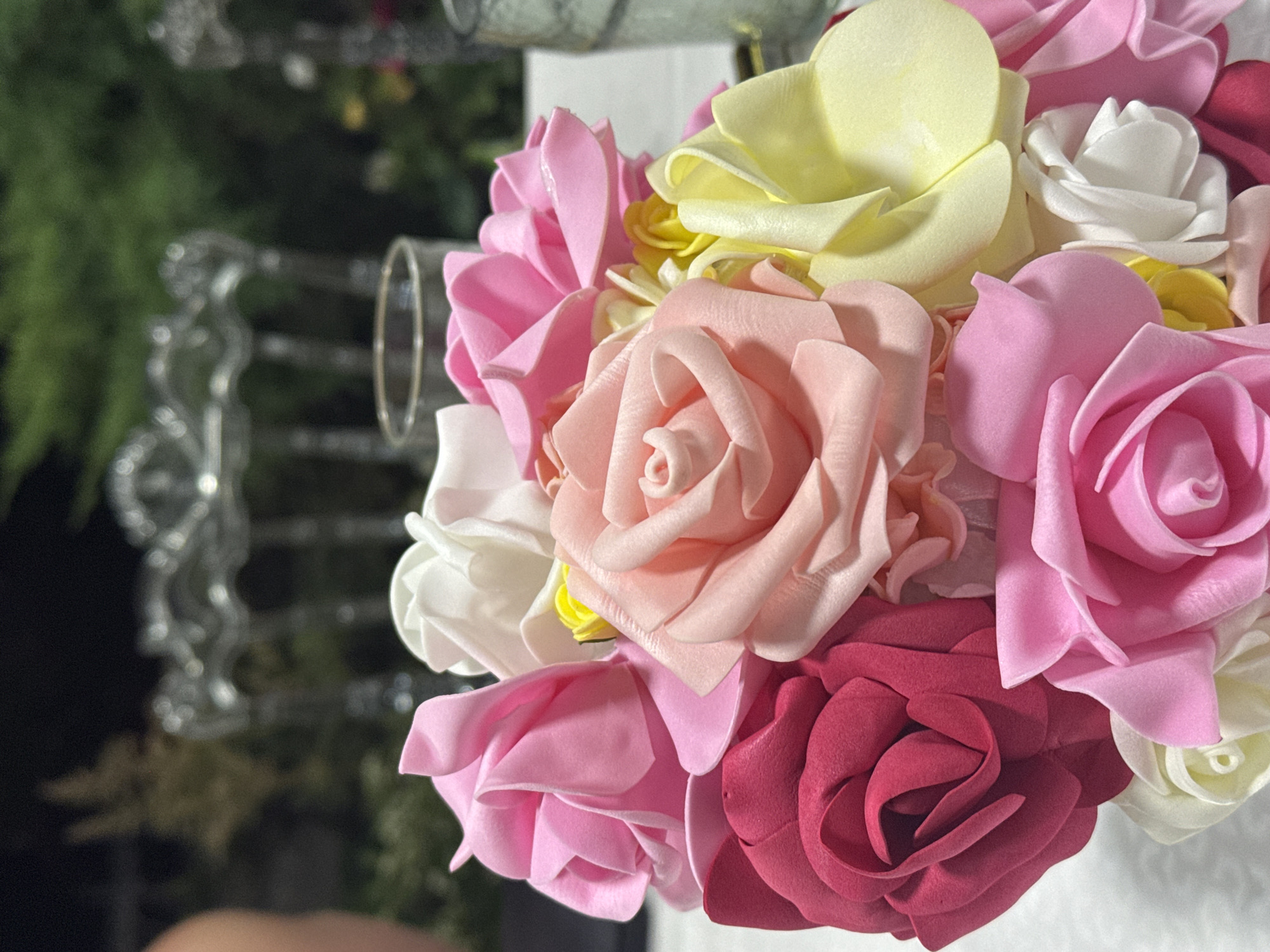 The image shows a close-up of a bouquet of artificial roses in various shades of pink, red, and yellow, with a few white roses interspersed. The roses appear to be made of foam or a similar soft material, with layered petals forming a delicate, textured surface. The bouquet is arranged in a way that creates a dense cluster of blooms, with some roses overlapping others. The background is blurred, suggesting an outdoor setting, possibly a garden or a reception area. Hints of greenery are visible, along with glimpses of ornate furniture, which could be chairs or a decorative screen. There are also hints of glassware and possibly candles, suggesting a celebratory or dining occasion. No people are clearly visible in the foreground or clearly identifiable. The overall impression is one of a decorative centerpiece, likely used for an event. The location is stated as Tash-Moinok, Kyrgyzstan.
