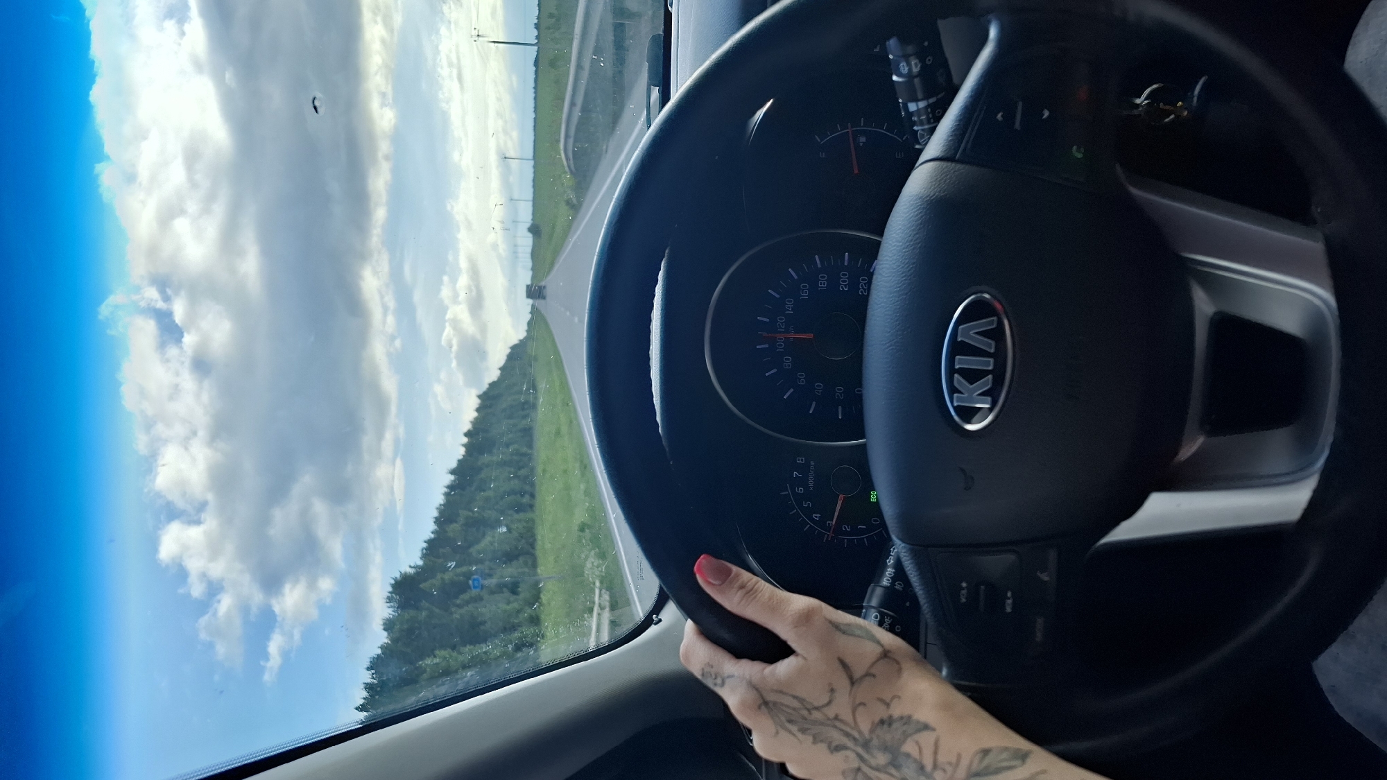 This is a photograph taken from inside a car, with the steering wheel and dashboard prominently featured. A woman's hand, with a pink painted nail and a tattoo on her forearm, is visible gripping the steering wheel. The car appears to be moving along a road, as indicated by the blur of the passing scenery and the visible speedometer on the dashboard which reads approximately 100 km/h. The road stretches ahead, flanked by lush green trees and fields, and the sky above is a bright blue with fluffy white clouds. The time of day seems to be daytime, likely late morning or afternoon, given the bright sunlight. The car is identified as a Kia by the logo on the steering wheel. The overall impression is one of a scenic drive on a pleasant day.