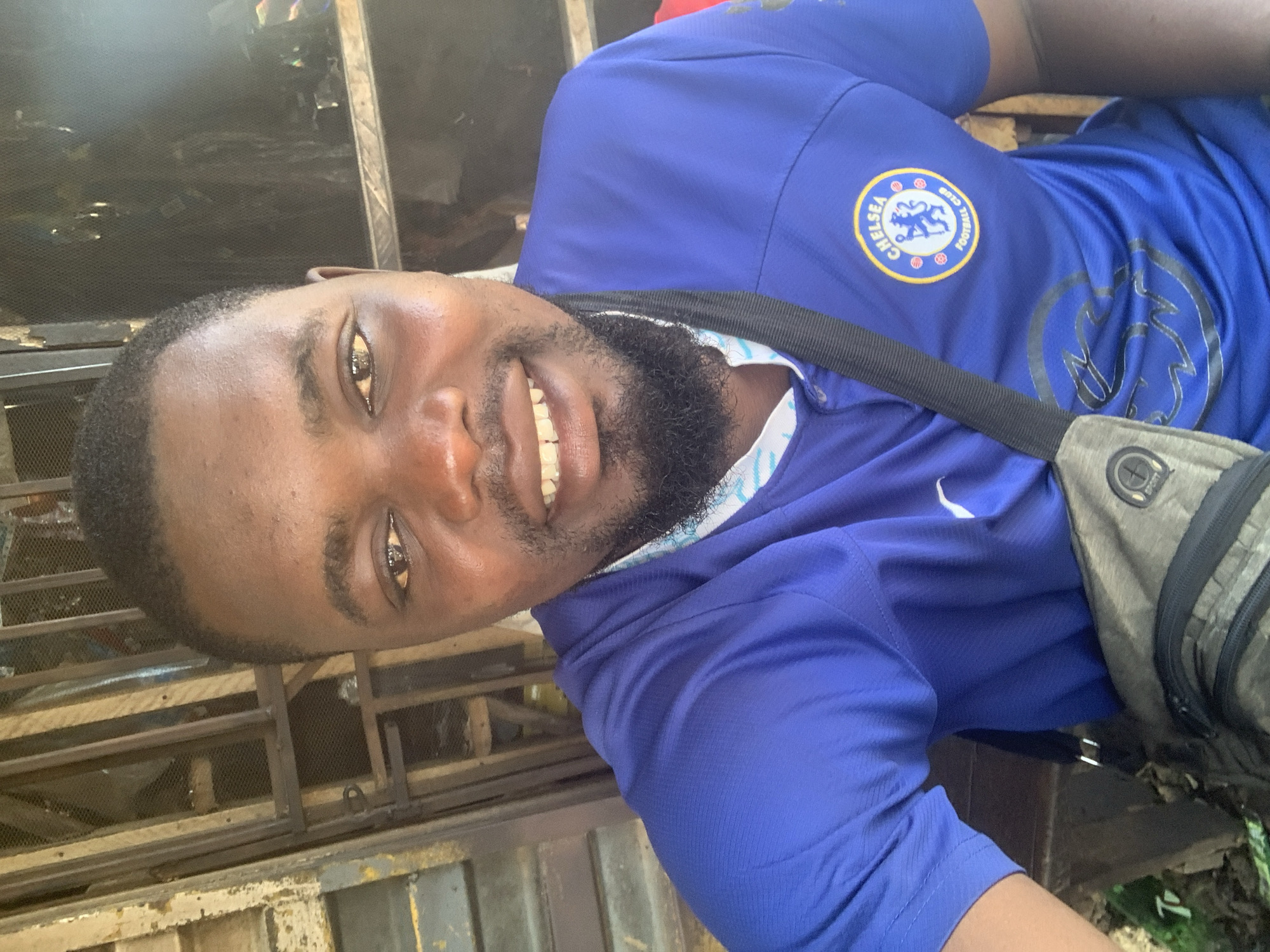 A man is taking a selfie outdoors. He is wearing a blue Chelsea Football Club jersey and a black strap is diagonally across his chest, from his right shoulder to his left hip. A grey crossbody bag is visible below the strap. His face is centered in the frame, with his mouth open in a smile, showing teeth. He has dark hair and a beard. The background appears to be a somewhat dilapidated outdoor area with metal bars and what looks like wood paneling. It is daytime, as indicated by the lighting. The location is the city of Shagamu, Nigeria.