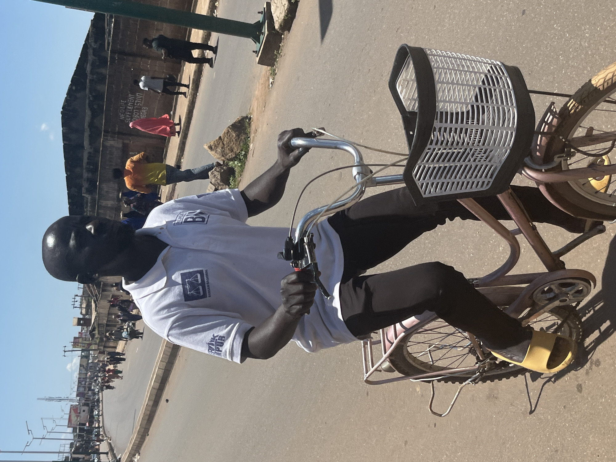 A Black man wearing a white polo shirt with a logo and black pants rides a bicycle with a front basket on a paved street in Jos, Nigeria. The man is facing forward, with his hands on the handlebars. He is wearing yellow slides on his feet. In the background, other people are walking on the sidewalk and in the street. There are buildings and a wall with graffiti in the background. The sky is clear and blue. The sun is shining brightly, casting shadows on the street. The overall scene depicts a typical day in a bustling city.