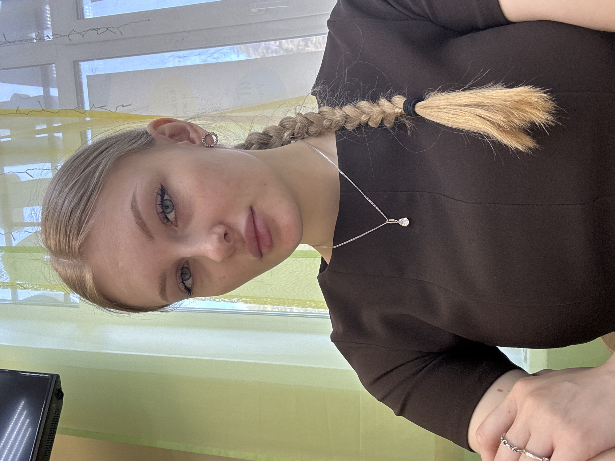 A young woman with fair skin, blond hair braided into a braid and secured with a black band, is posing. She is wearing a dark brown shirt and a silver necklace with a pendant. She has a silver ring on her right hand. The background shows a window with translucent green curtains. The scene appears to be indoors. It is not possible to identify the location as the city of Perm, Russia, from this image.