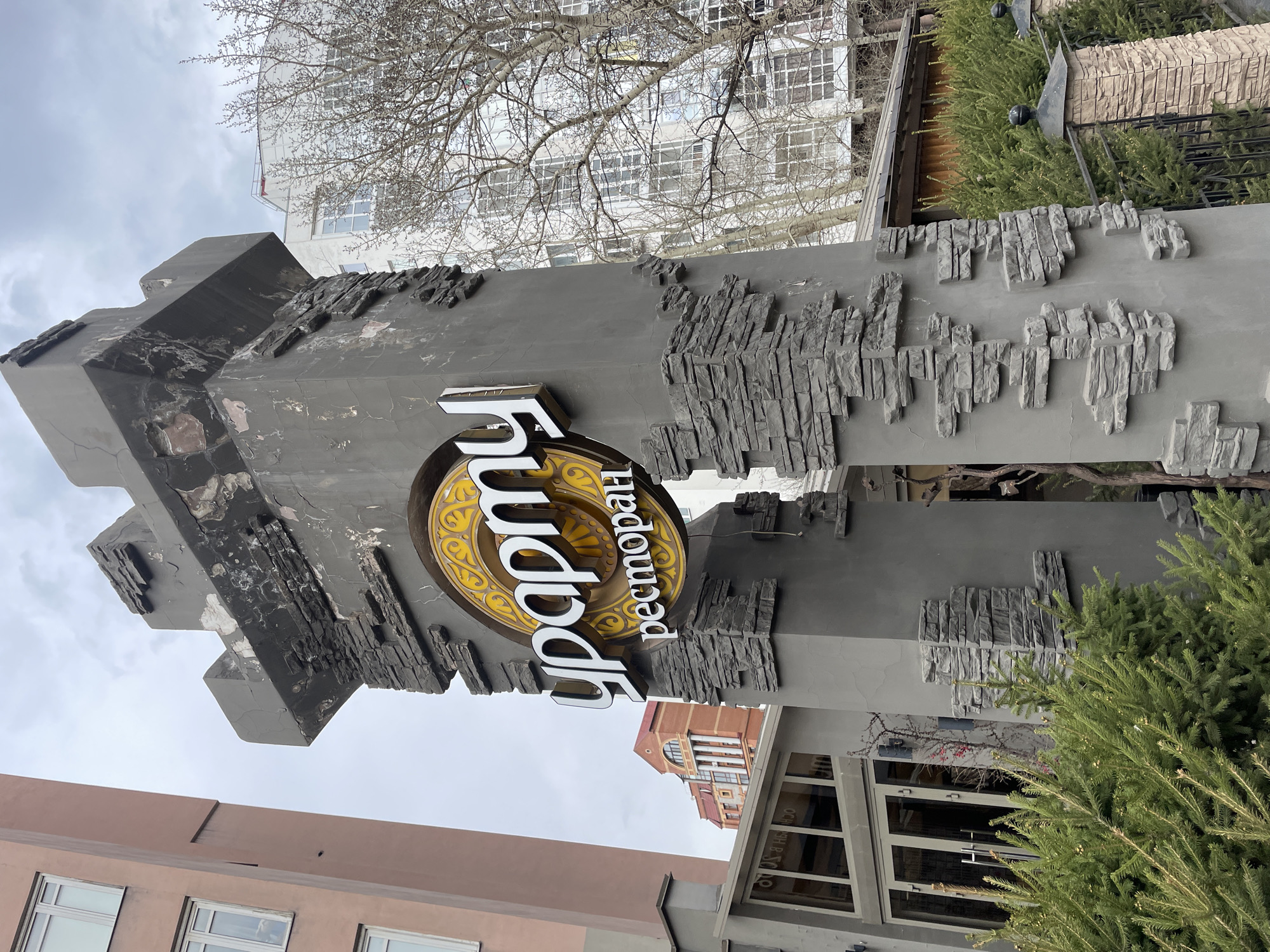 The image shows the exterior signage for the "Урфин" restaurant, located in Krasnoyarsk, Russia. The sign is a large, gray, stone-textured pillar with an elaborate design. A circular emblem with gold-colored ornamental patterns surrounds the white, Cyrillic letters spelling "Урфин." Beneath the emblem is the word "ресторан" (restaurant) written vertically. The pillar has a somewhat dilapidated appearance.
In the background, there are several buildings, including a white one with numerous windows and a red brick building with classical architectural elements. Bare tree branches are visible against the overcast sky. Green coniferous plants are placed on the right side of the image, adjacent to the pillar.