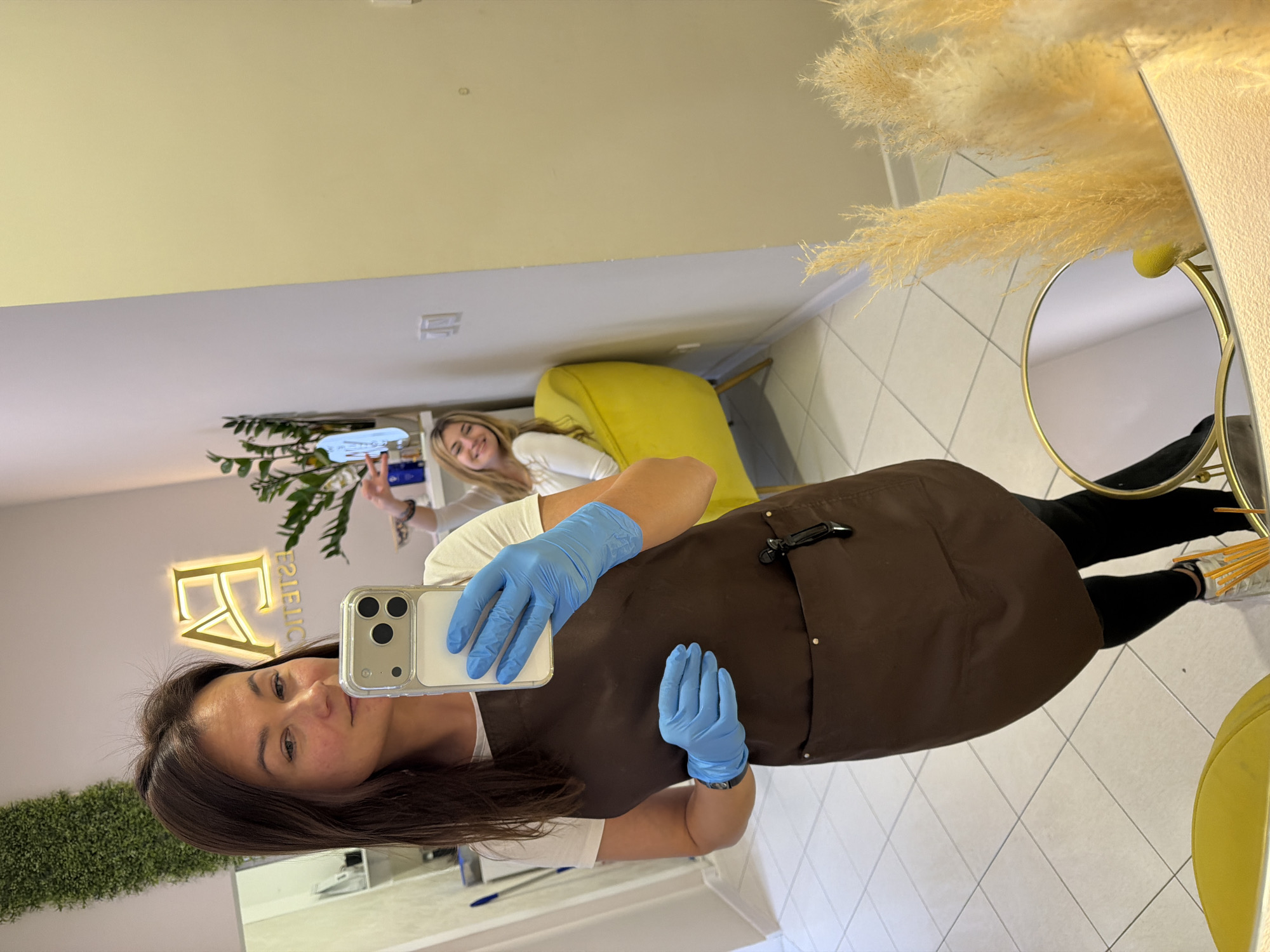 This image is a mirror selfie taken by a woman in what appears to be a beauty salon or aesthetic studio located in Budva, Montenegro. The photo is rotated 90 degrees clockwise.
The main subject, positioned in the foreground, has long dark brown hair and is looking directly at her smartphone, which she holds up to capture the mirror reflection. She is wearing a white t-shirt, a dark brown apron or smock, and light blue examination gloves on both hands, indicative of a professional setting. Her expression is neutral and pleasant.
Reflected in the background is a second woman with blonde hair, seated on a vibrant yellow chair. She is smiling broadly and making a peace sign with her right hand.
The indoor setting features light-colored walls and a light-tiled floor with a diamond pattern. Decorative elements include a large arrangement of fluffy, beige pampas grass on the right, and green leafy plants, one on a shelf and another as a faux green wall texture on the left. A prominent, illuminated golden sign is mounted on the wall behind the main subject's reflection. The mirrored text on the sign reads "FA ESTETIC." A smaller, circular mirror with a gold frame is also visible in the background. The space is well-lit, suggesting either daytime or bright artificial lighting. Both individuals appear to be in good spirits, conveying a friendly and professional atmosphere.