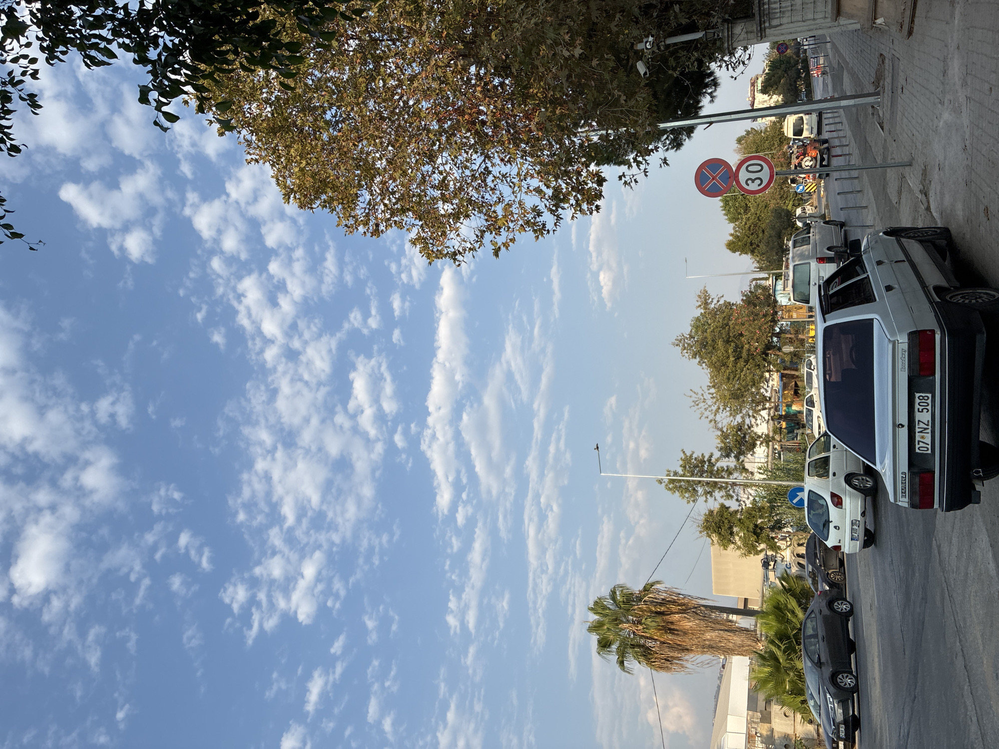 The image captures a street scene in Alanya, Turkey, under a bright blue sky filled with scattered white clouds. On the right side of the road, a tree with autumnal foliage is prominent, its branches partially obscuring the sky. Adjacent to the tree is a sidewalk with a stone wall and a lamppost. Several traffic signs are visible on poles along the sidewalk, including a red and blue circular sign with a cross and a circular sign indicating a speed limit of 30. Security cameras are also mounted on a pole near the signs.
The street is paved and appears to be a two-way road with multiple cars parked or driving. A light-colored sedan is parked in the foreground on the right side of the street, facing away from the viewer. Further down the street, other vehicles are visible, including a white hatchback, a dark-colored sedan, and several vans. A palm tree stands on the left side of the road, with a building and some industrial structures visible in the background. The overall impression is a typical urban streetscape on a clear day.