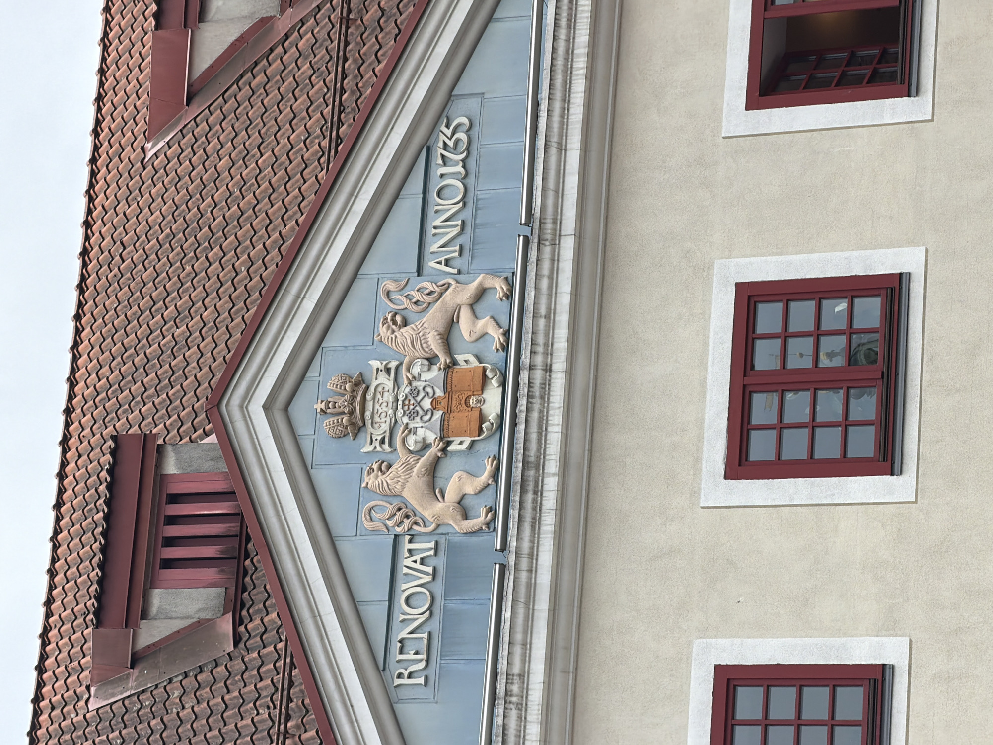 The image shows the facade of a historical building, likely a public institution or a well-preserved residence, in the city of Riga, Latvia. The building features a red tiled roof with a dormer window, and a decorative pediment above the upper floor. The pediment is adorned with a crest featuring two lions, a crown, and the inscription "RENOVAT ANNO 1735". Below the pediment, there are several windows with red frames. The overall architectural style suggests a building with a significant history, possibly dating back to the 18th century. The area appears to be a well-maintained urban environment.