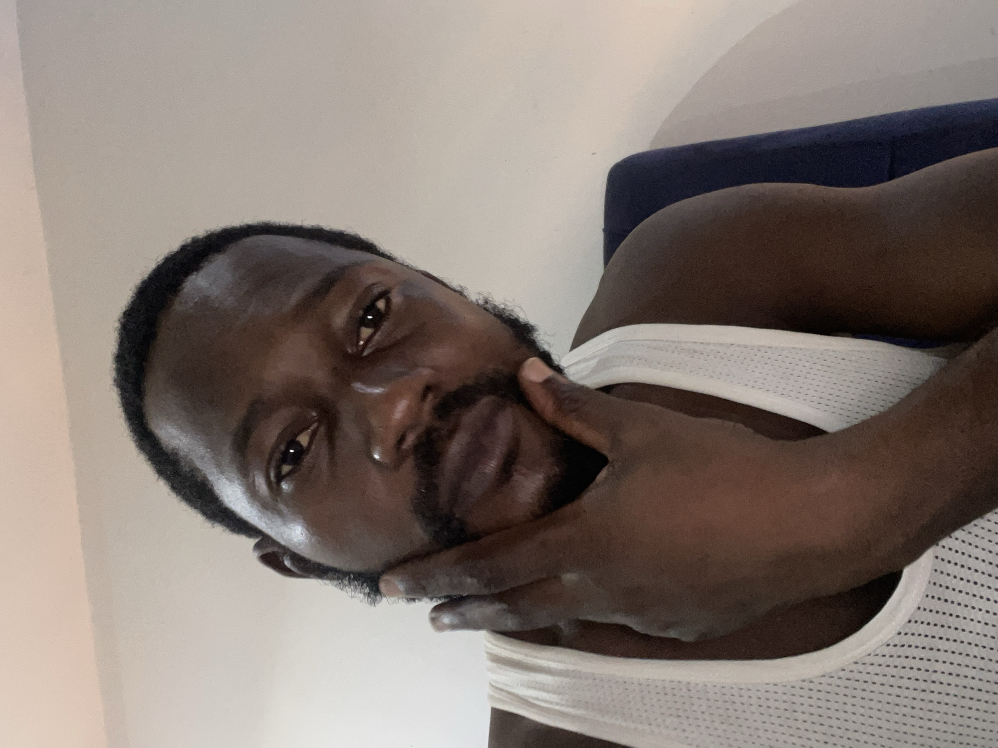 The image depicts a man with dark skin and a beard. He is wearing a white tank top and lying on a blue cushion. He is touching his face with his right hand. The location in Abuja, Nigeria, is not discernible from the image.