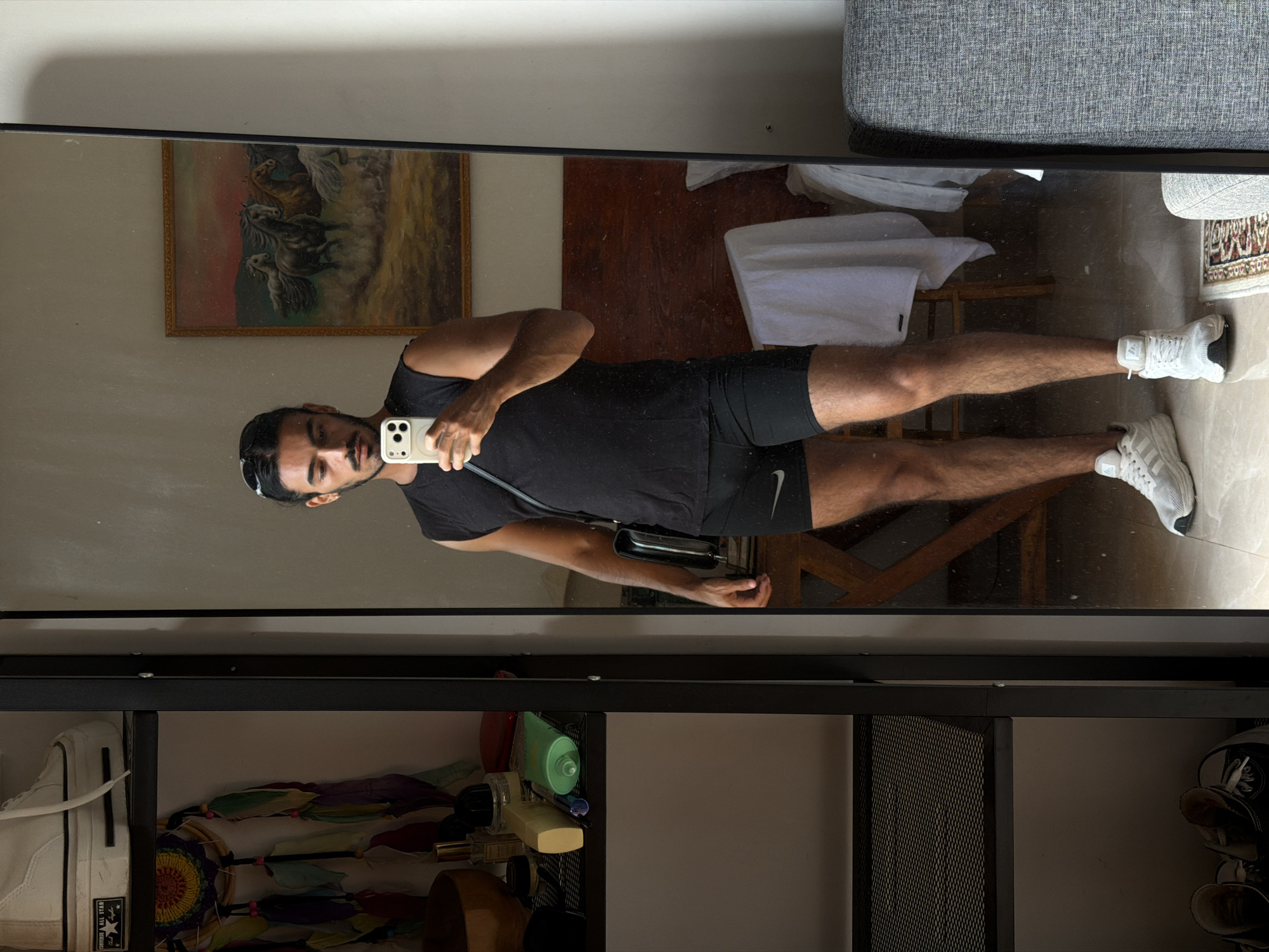The image shows a man taking a selfie in a full-length mirror. He is wearing a black tank top and black shorts with a white Nike logo, and white athletic shoes. He is holding a smartphone in his left hand. A black crossbody bag is slung over his shoulder. The mirror reflects a room with a painting of horses hanging on the wall. There is a bed with white linens and a wooden chair in the background. A gray sofa is partially visible in the top right corner. Below the mirror, a black metal shelf unit contains various items, including shoes, bottles, and a dreamcatcher. The location is North Kuta, Indonesia.