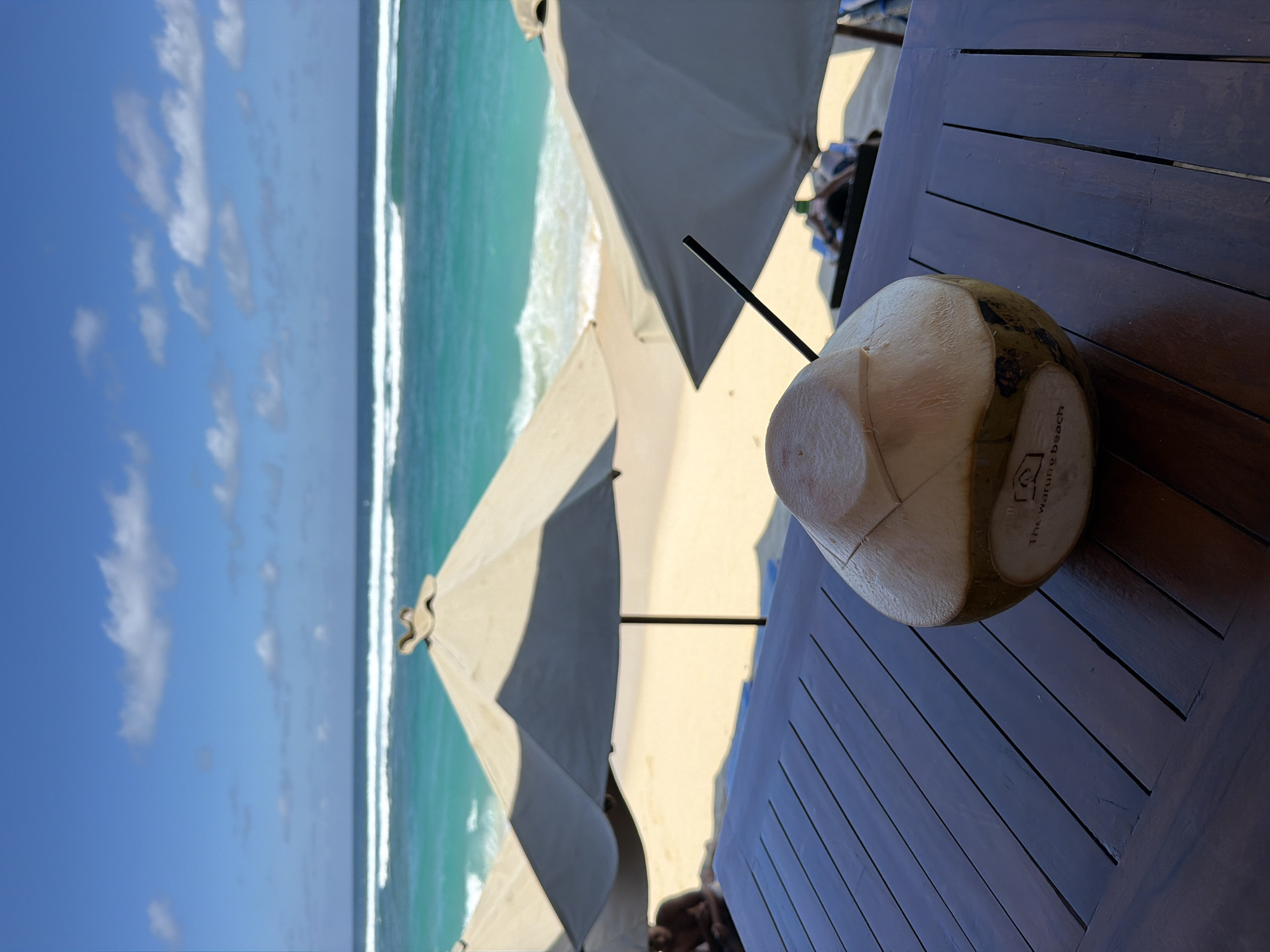 The image, presented initially in a vertical orientation, captures a serene tropical beach scene from a shaded, elevated vantage point. In the immediate foreground, a dark, slatted wooden table prominently displays a fresh coconut drink with a black straw. The side of the coconut bears visible text: "The warung beach" and a small, distinct logo that appears to be a stylized hut or house.
Beyond the table, the bright sandy beach is partially visible, featuring several large, light-colored beach umbrellas casting strong shadows, indicative of bright daylight conditions, likely midday or afternoon. The vibrant turquoise-green ocean stretches across the midground, with gentle white waves breaking along the shore. The background is dominated by a clear blue sky dotted with scattered white clouds. No specific individuals are clearly discernible, but the setup suggests a relaxing environment, possibly at a beach club or resort. The visible text on the coconut strongly hints at the specific location being "The Warung Beach," situated in Kuta Selatan, Indonesia.