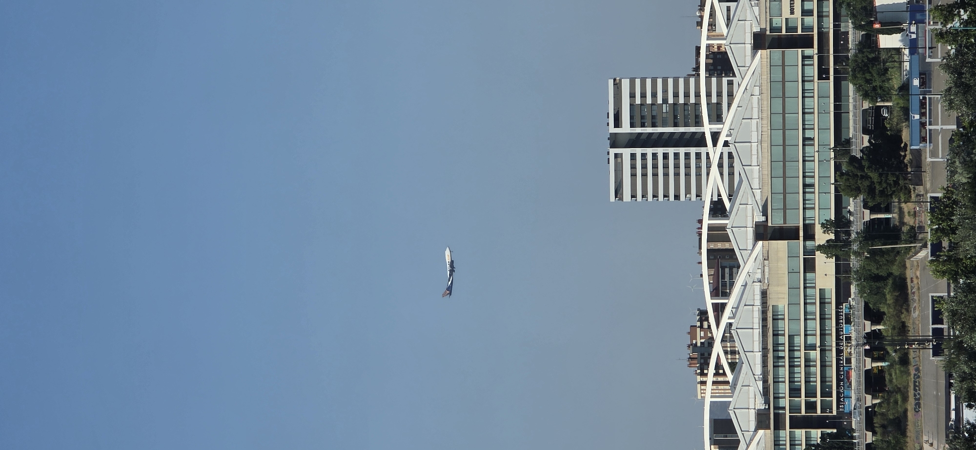 The image, likely taken in Zaragoza, Spain, depicts a clear blue sky with a single, small airplane visible in the distance. The airplane appears to be white with blue and dark accents and has a flag or banner trailing behind it. The majority of the image is dominated by the sky, suggesting an outdoor setting with good visibility.
To the right of the frame, a modern architectural complex rises, featuring several buildings with glass facades and distinct geometric designs. These buildings are flanked by trees and a road, suggesting an urban environment. One of the buildings has text visible on its facade: "ESTACION CENTRAL DE AUTOBUSES," which translates to "Central Bus Station." The orientation of the image is rotated 90 degrees counter-clockwise. The sunlight suggests it is daytime, and the clear sky indicates fair weather. The overall impression is one of a bright, possibly sunny day in a city with contemporary architecture and functional infrastructure.