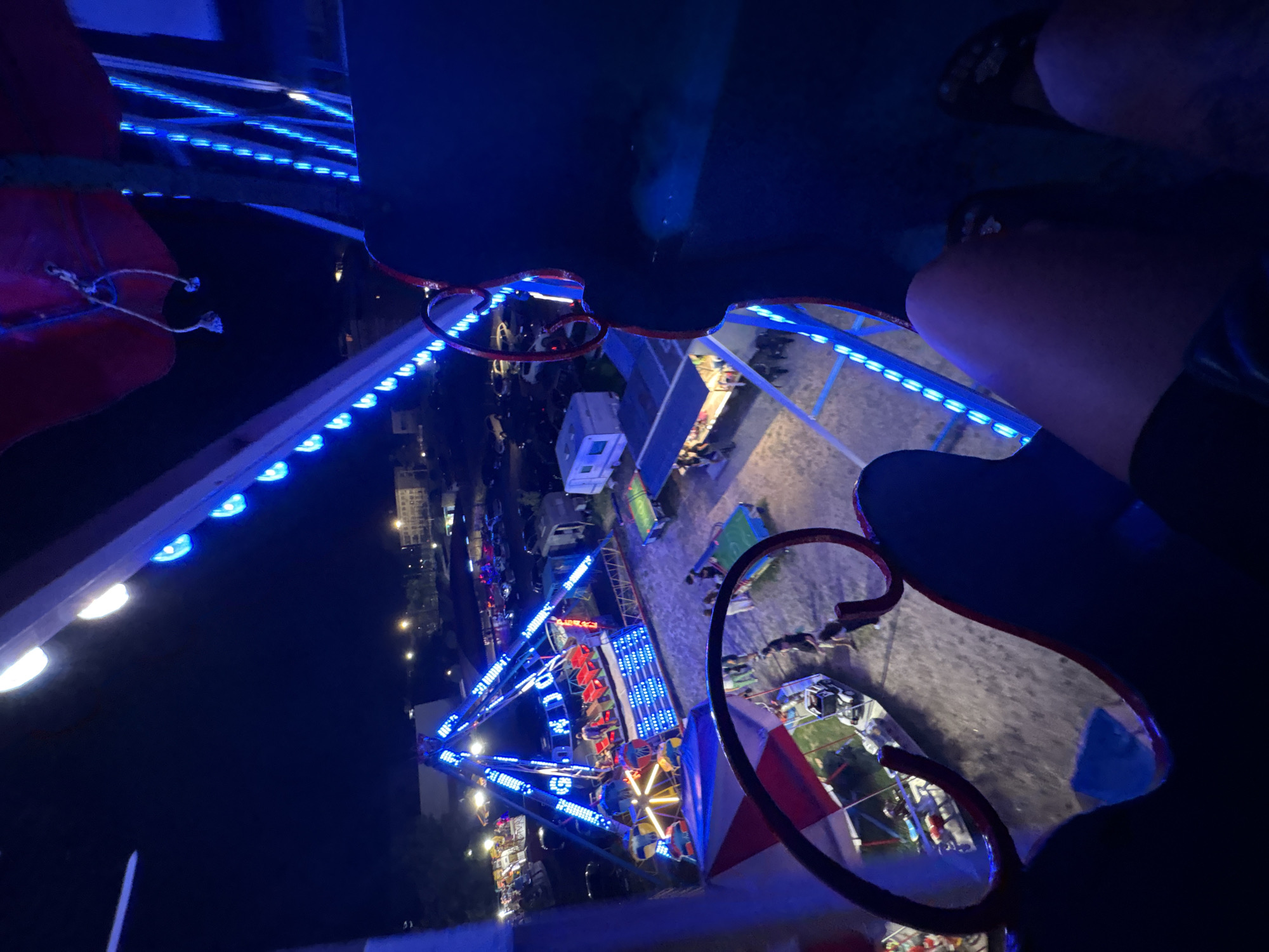 The image depicts a nighttime scene at a fairground in Primorsko, Bulgaria. The perspective is from a ride, likely a Ferris wheel, looking down and outward. The foreground features the dark interior of the ride's cabin or seating area, with parts of a person's legs and shoes visible. Bright blue LED lights line the structure of the ride and other attractions in the background, illuminating the fairground at night.
Dominating the mid-ground is a large amusement ride, possibly a pendulum or drop tower, adorned with numerous blue lights. Several people are seen on and around this ride. Beyond this main attraction, a variety of other fairground elements are visible, including smaller rides, food stalls or game booths with awnings, and parked vehicles, suggesting a bustling event. In the distance, buildings and streetlights indicate the presence of the town of Primorsko. The ground appears to be a mixture of sand and gravel, typical of outdoor fairgrounds. The overall atmosphere is festive and energetic, enhanced by the vibrant lights against the dark night sky.